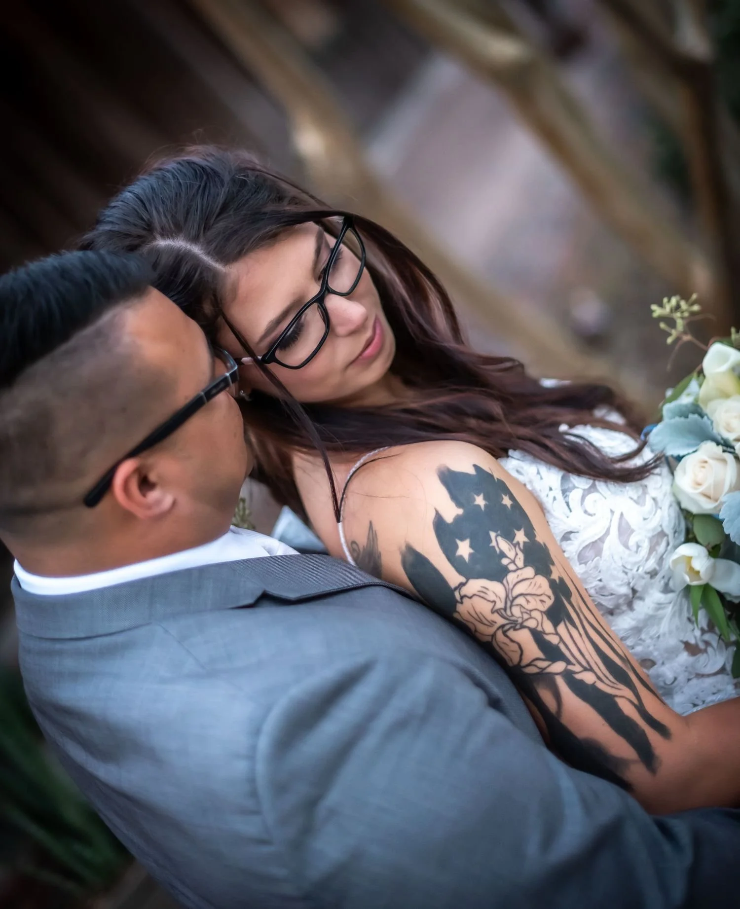 A photo taken by Trisha Disney-Linder features a woman with glasses and a tattoo on her arm is leaning on a man in a gray suit at a wedding or formal event, with floral arrangements nearby.
