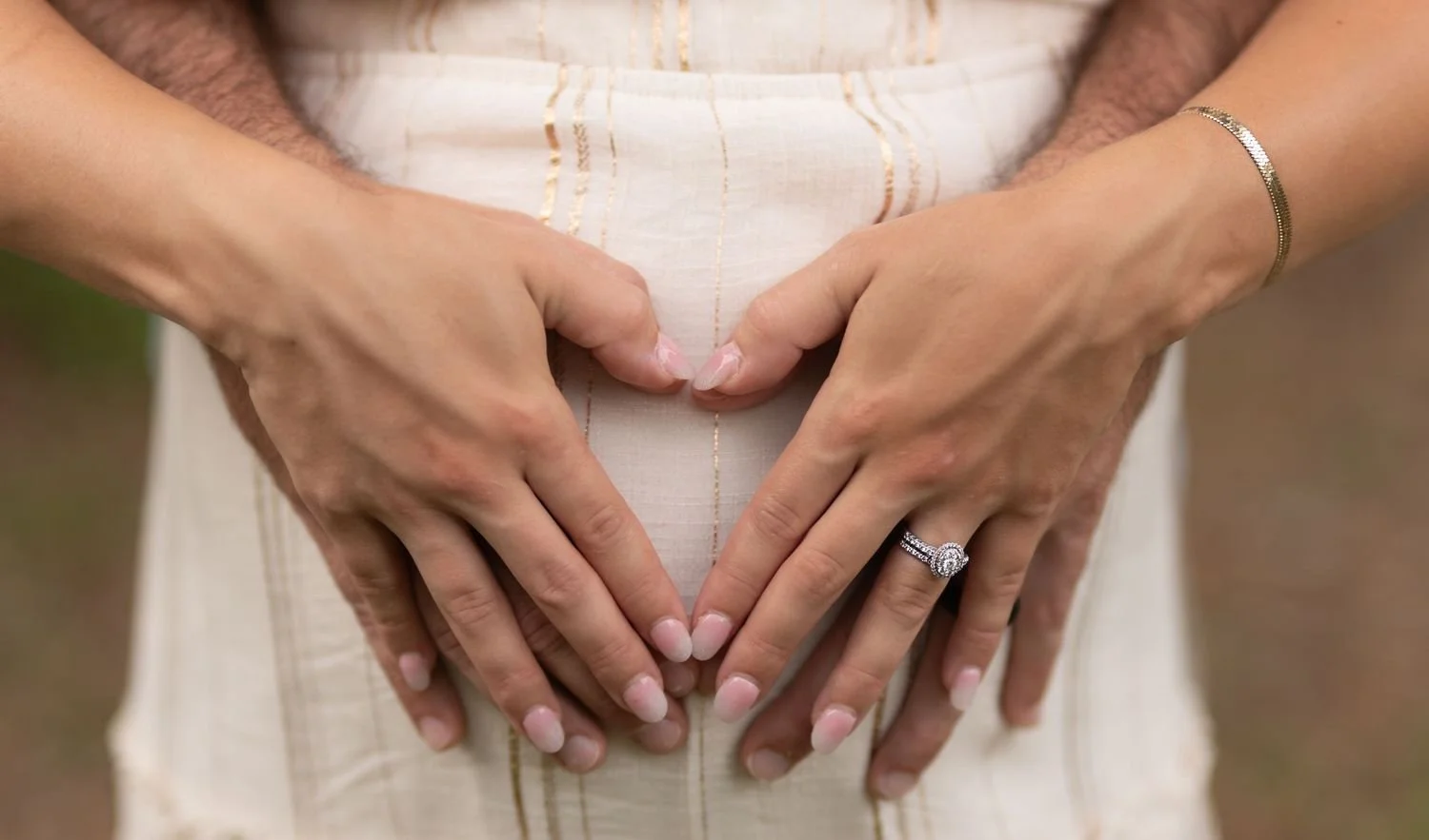 Close-up of a pregnant woman with her partner's hands on her belly, forming a heart shape, with an engagement ring visible.