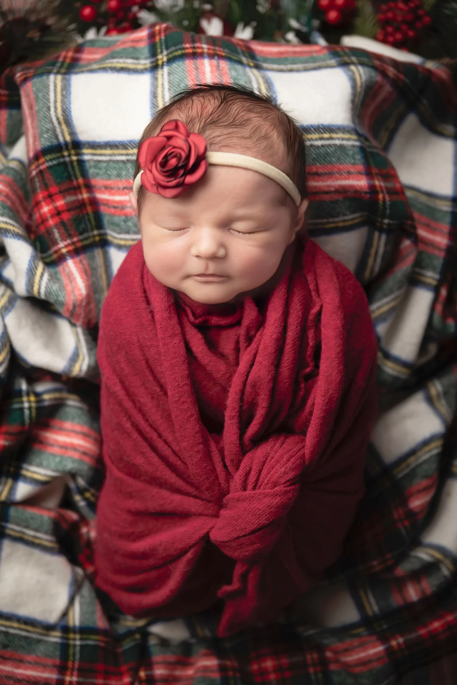 A sleeping newborn baby wrapped in a red blanket, wearing a headband with a red flower, lying on a plaid blanket.