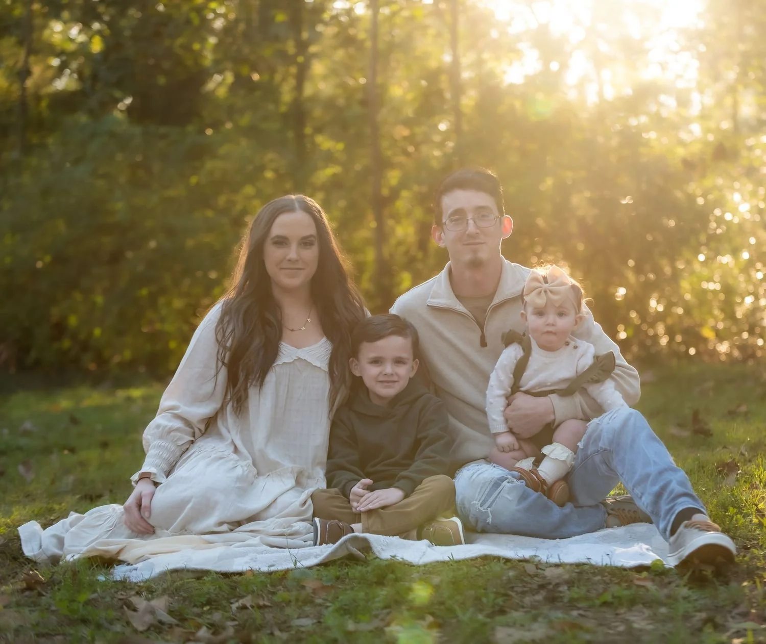 Family of four sitting on a blanket outdoors during sunset, surrounded by trees.