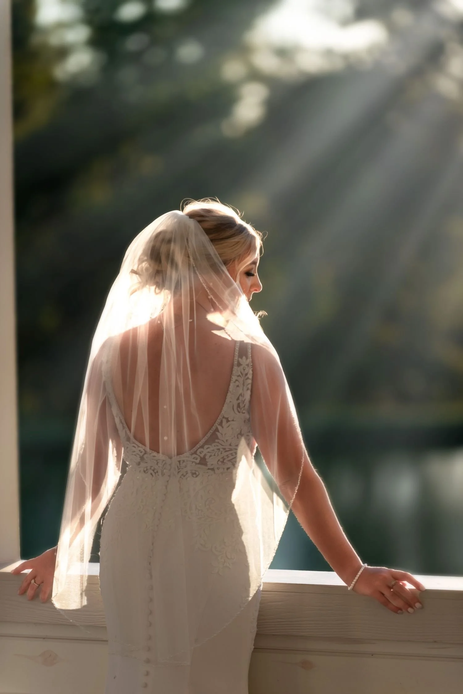 A photo taken by Trisha Disney-Linder features a bride in a lace wedding dress with a veil standing by a window, gazing at sunlight streaming through trees outside.
