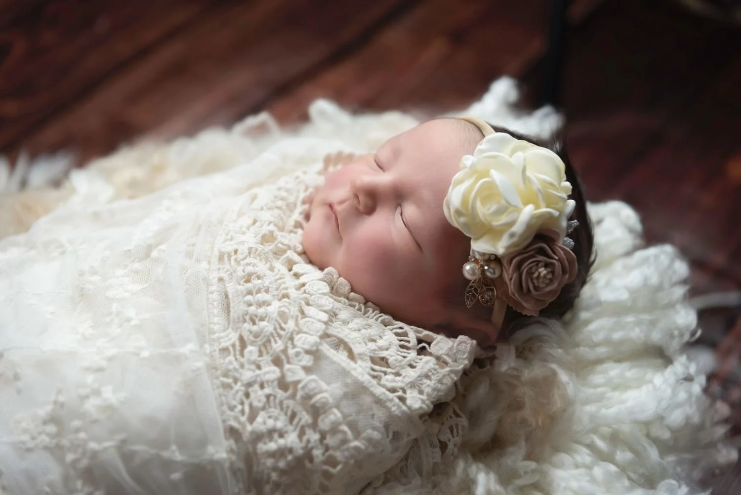 A sleeping baby girl wearing a lace dress and floral headband, lying on a fluffy white blanket and wooden surface.