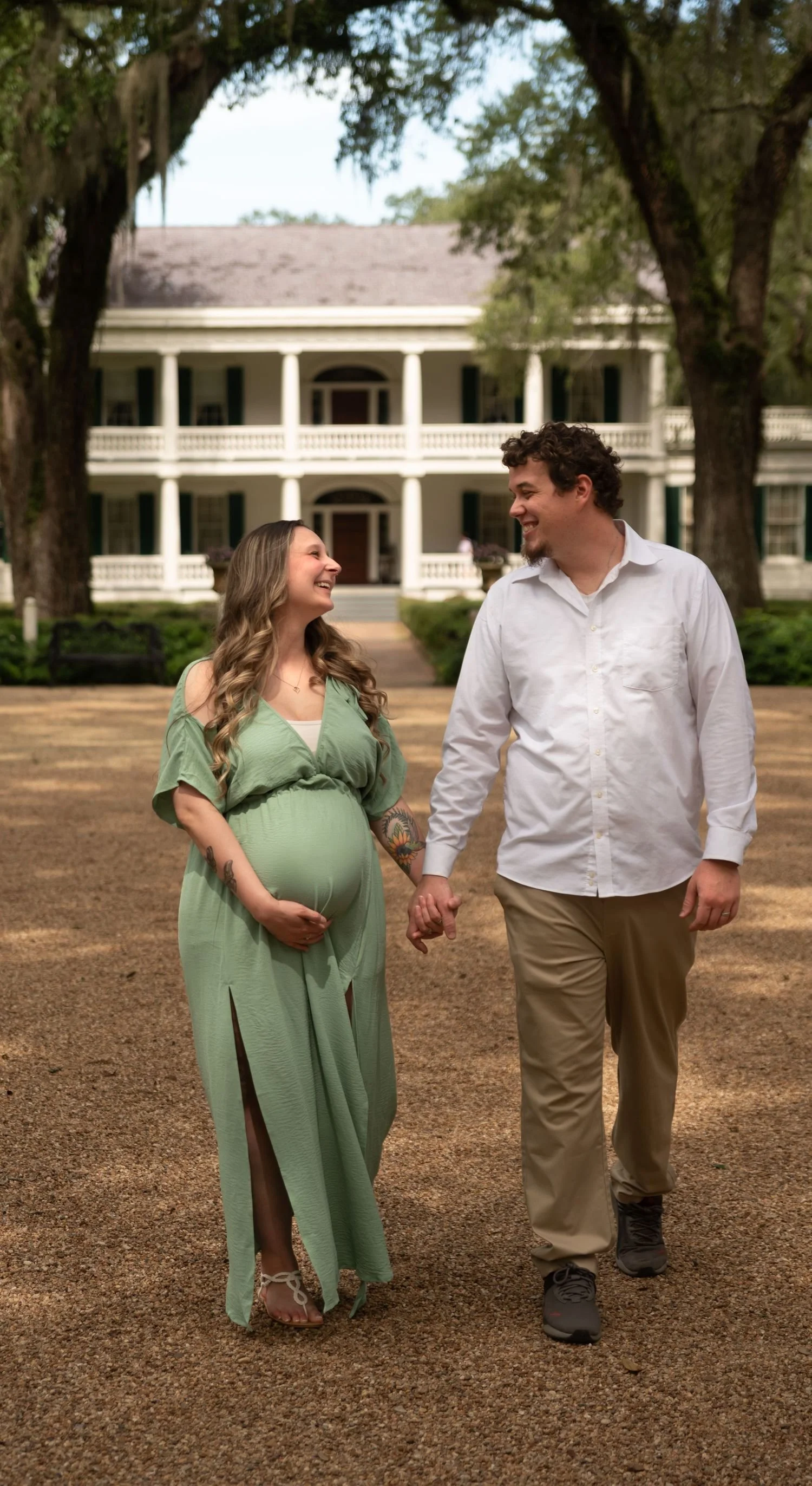 Pregnant woman in green dress holding her belly, smiling at man dressed in white shirt and khaki pants, holding hands, walking together outdoors in front of a large house with a porch and trees.