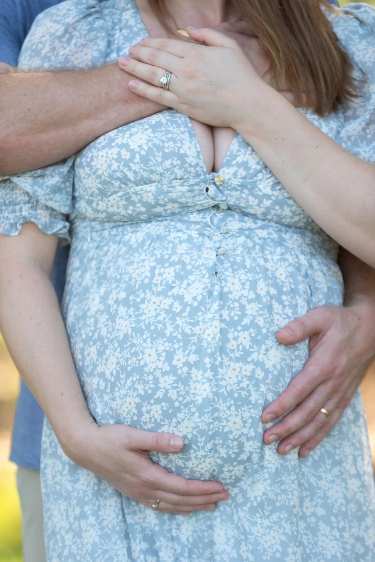 A pregnant woman wearing a floral dress with a man standing behind her, both holding her belly with their hands, outdoors.