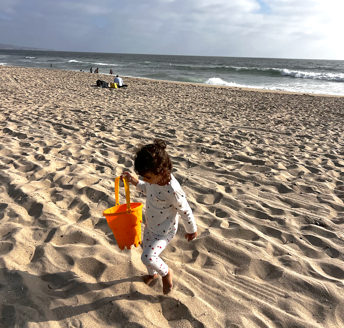 A young girl walking on the sandy beach with a yellow bucket, with the ocean and other beachgoers in the background in Manhattan Beach, CA.