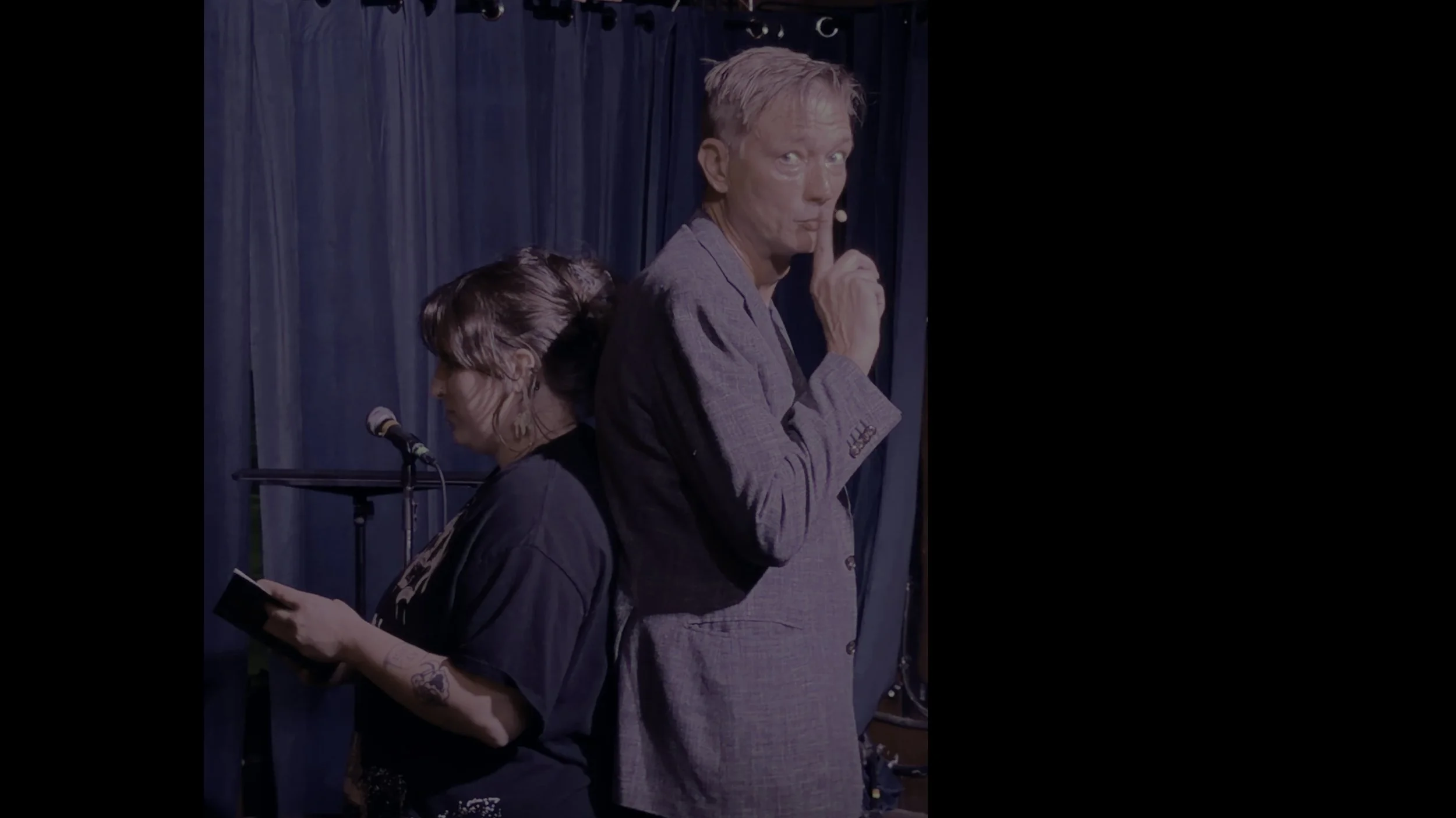 A man with blonde hair making a shushing gesture with his finger on his lips, standing on stage with a woman who is looking down, holding a book