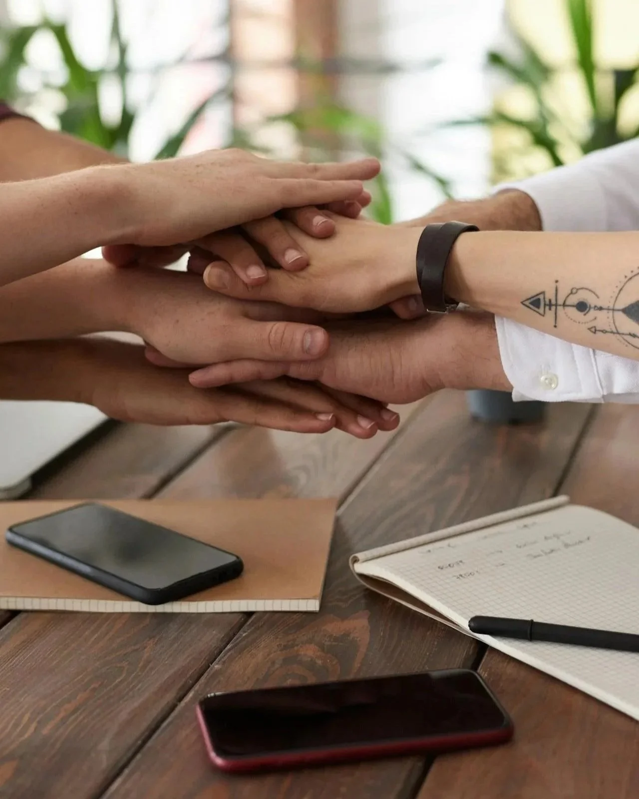 Flere hender som holder hverandre i en gruppe på et bord med notatbøker og mobiltelefoner, utstråler samarbeid og fellesskap.