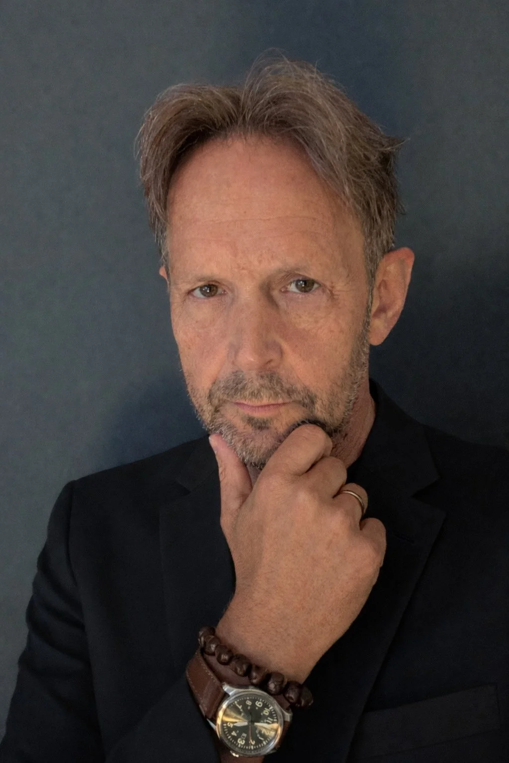 Porträtt av en man med lätt skägg, grått hår och en svart kostym, som reflekterar ett tänkande uttryck medan han håller sin käke med handen, med en mörk bakgrund.