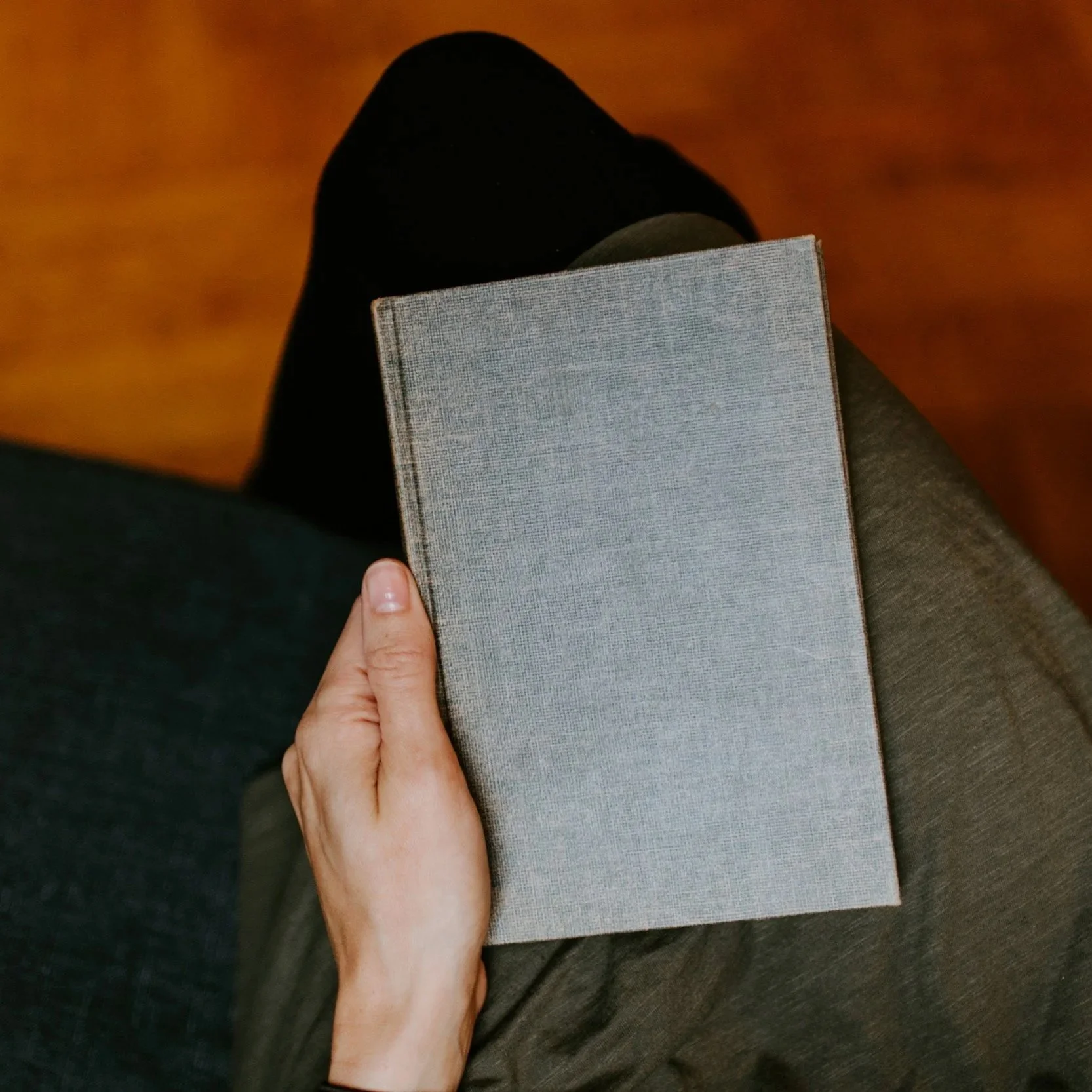 Person sitting with a gray book resting on their lap, wearing dark pants and a black top.