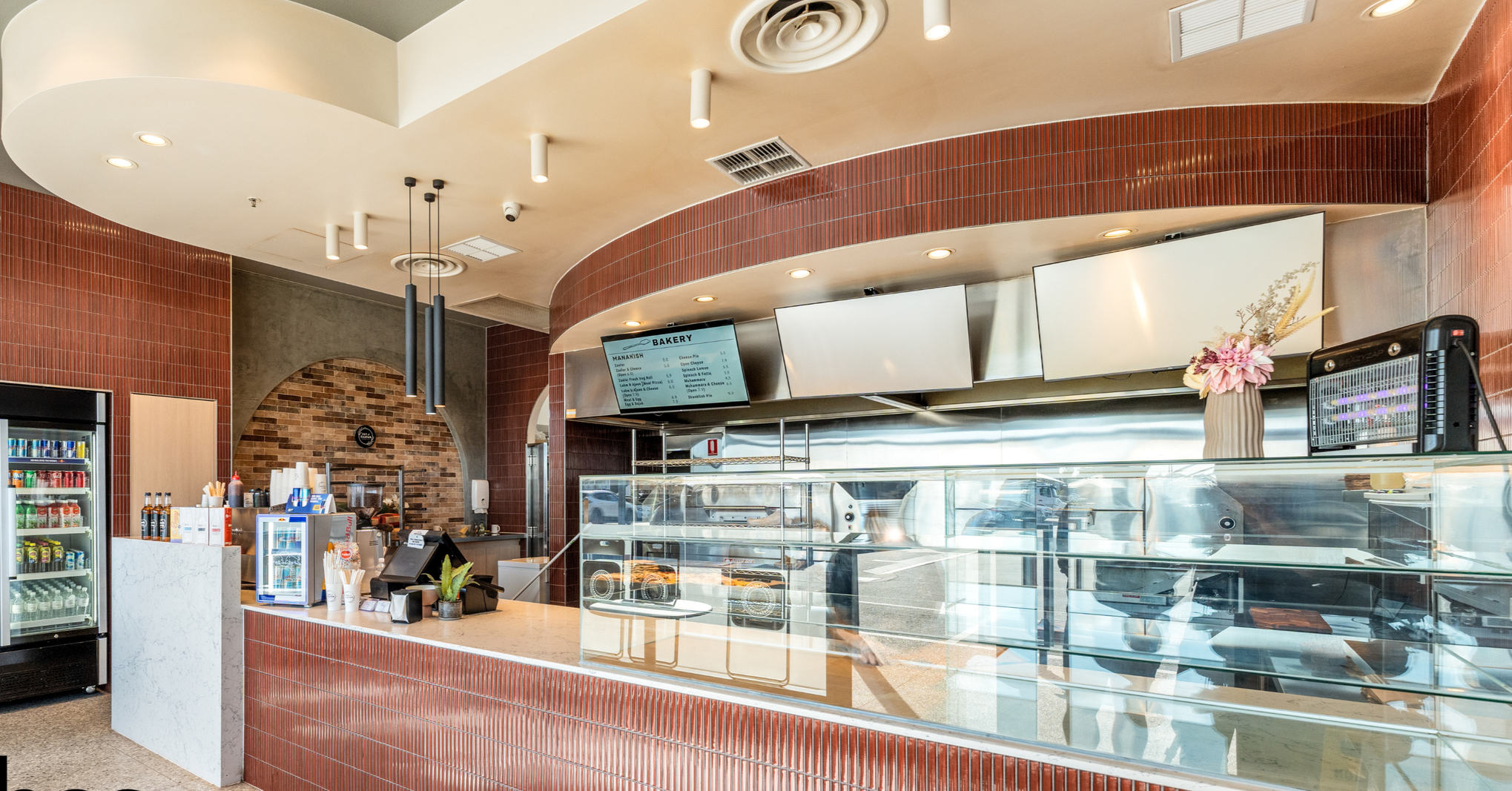 Inside a bakery with a glass display case, digital menu screens, a refrigerator, and a counter with a cash register, decorated with a vase of pink flowers.