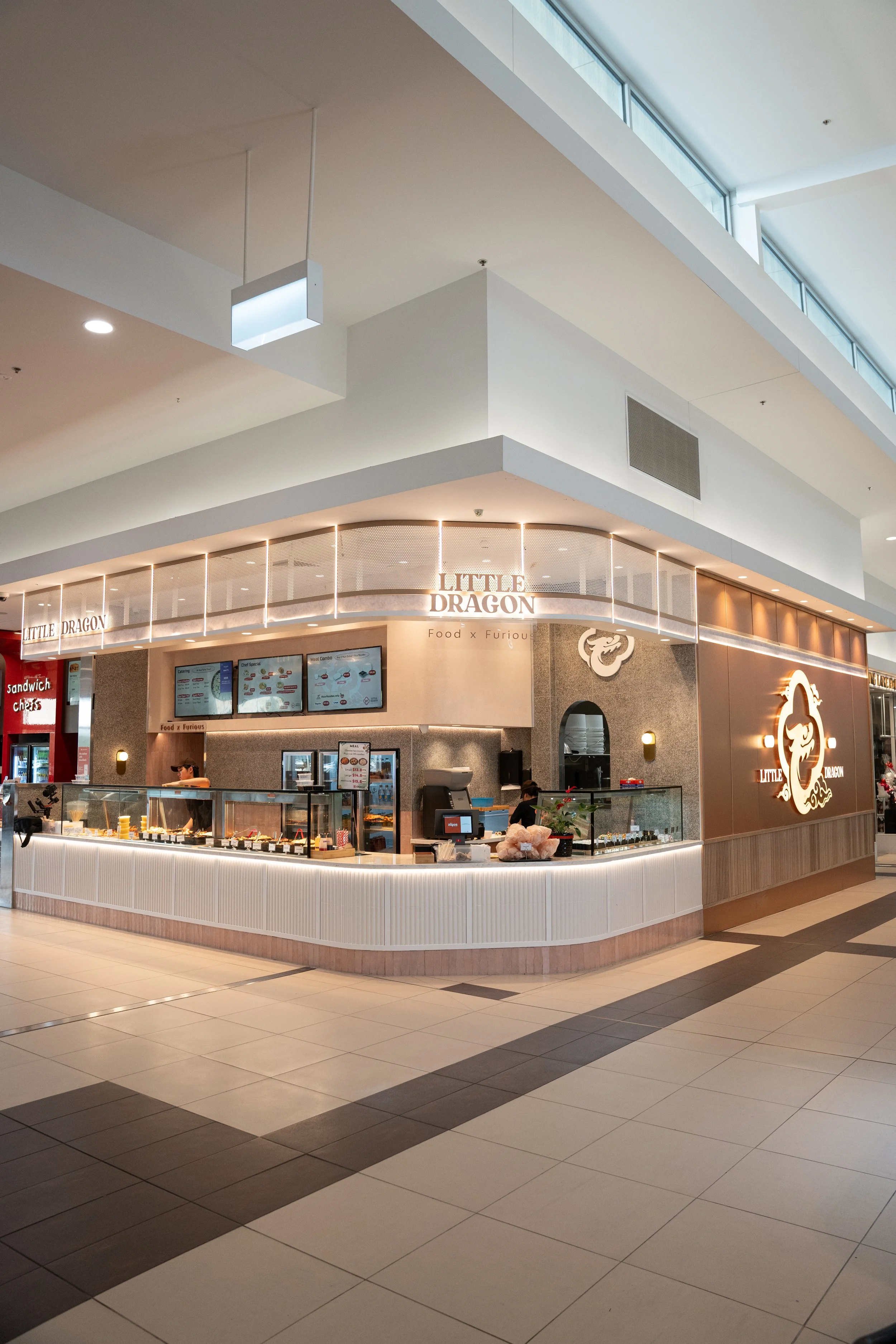 A food court kiosk named Little Dragon with a modern design, illuminated signage, digital menu screens, and a display case of food items, located in a mall.