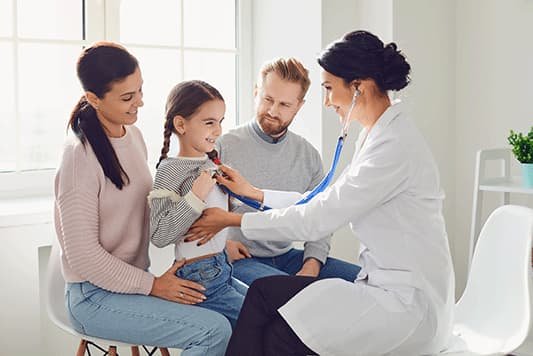 Doctor examining a young girl with a stethoscope while her mother and father look on in a bright, modern clinic.