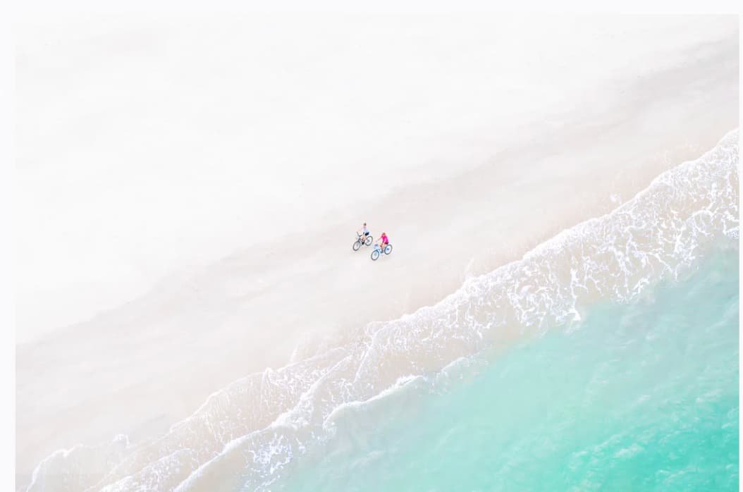 Two people riding bicycles along a beach shoreline with waves washing up on the sand.