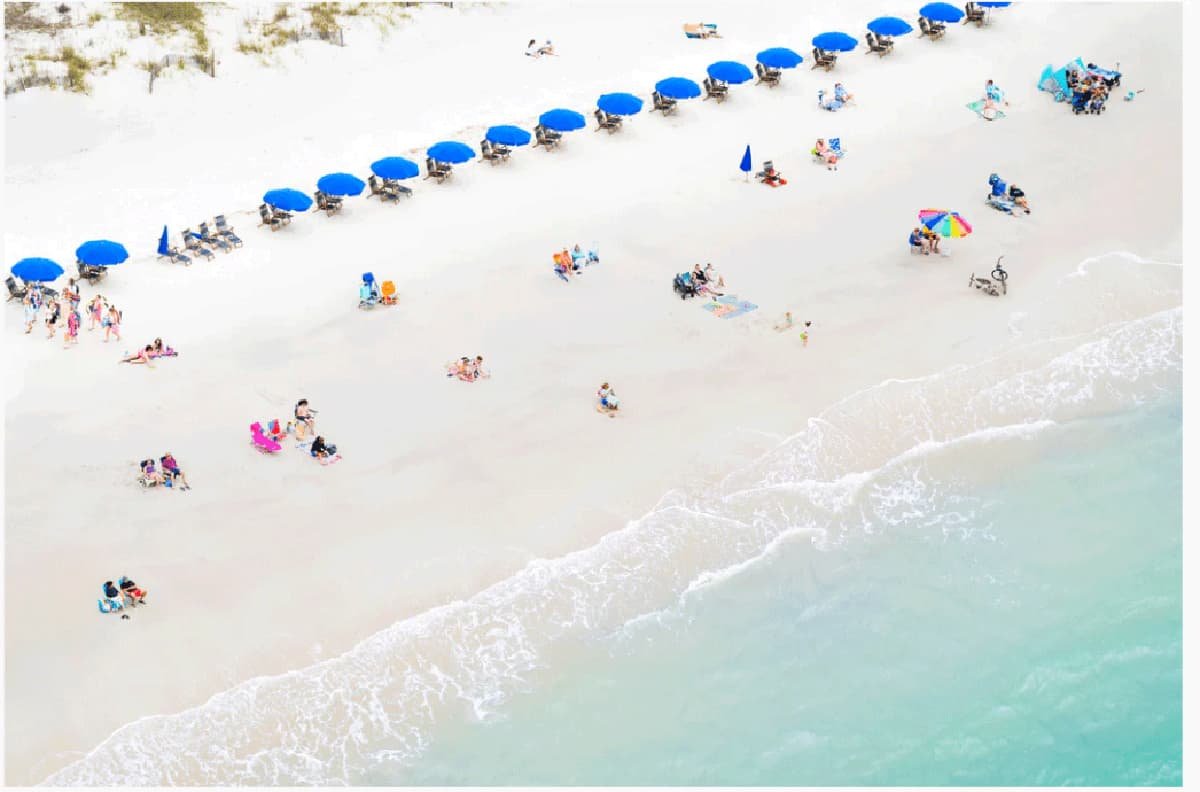 Beach scene with blue umbrellas, lounge chairs, and people sunbathing, swimming, and playing on white sand and near the ocean.