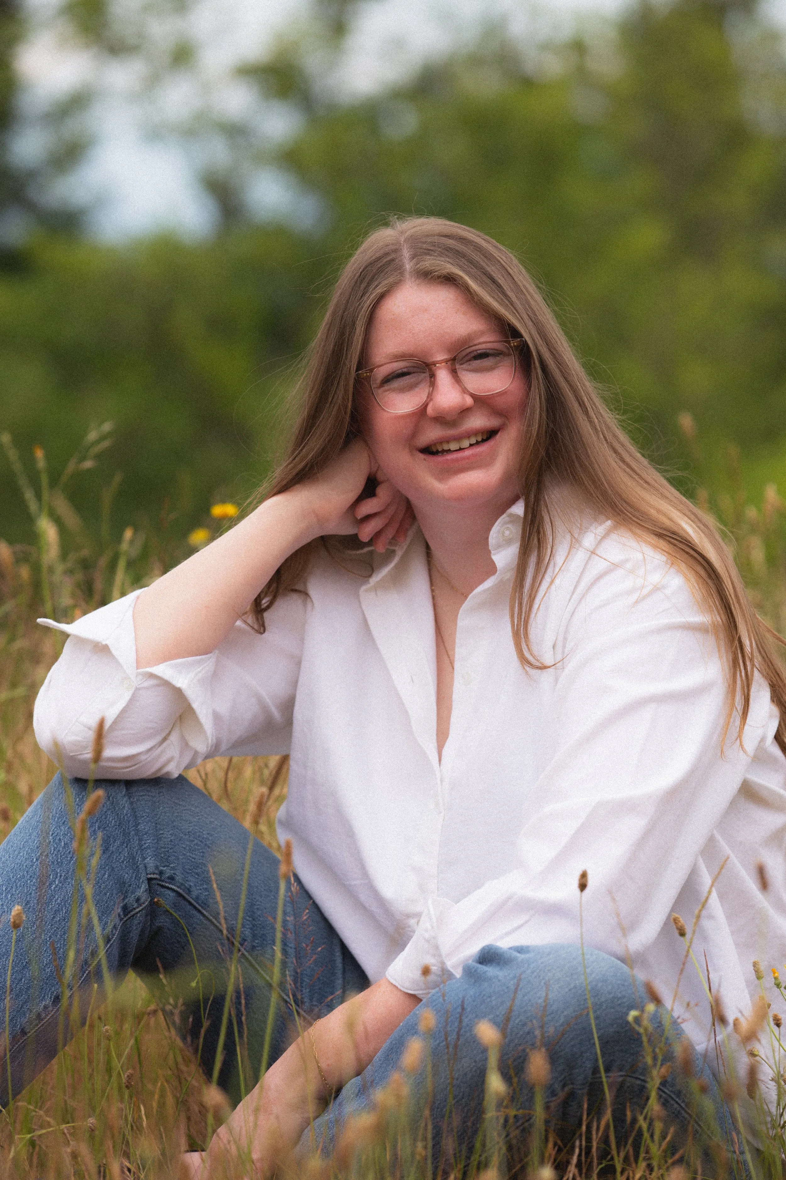 Young woman with long hair and glasses sitting in a grassy field, smiling at the camera, wearing a white shirt and blue jeans.