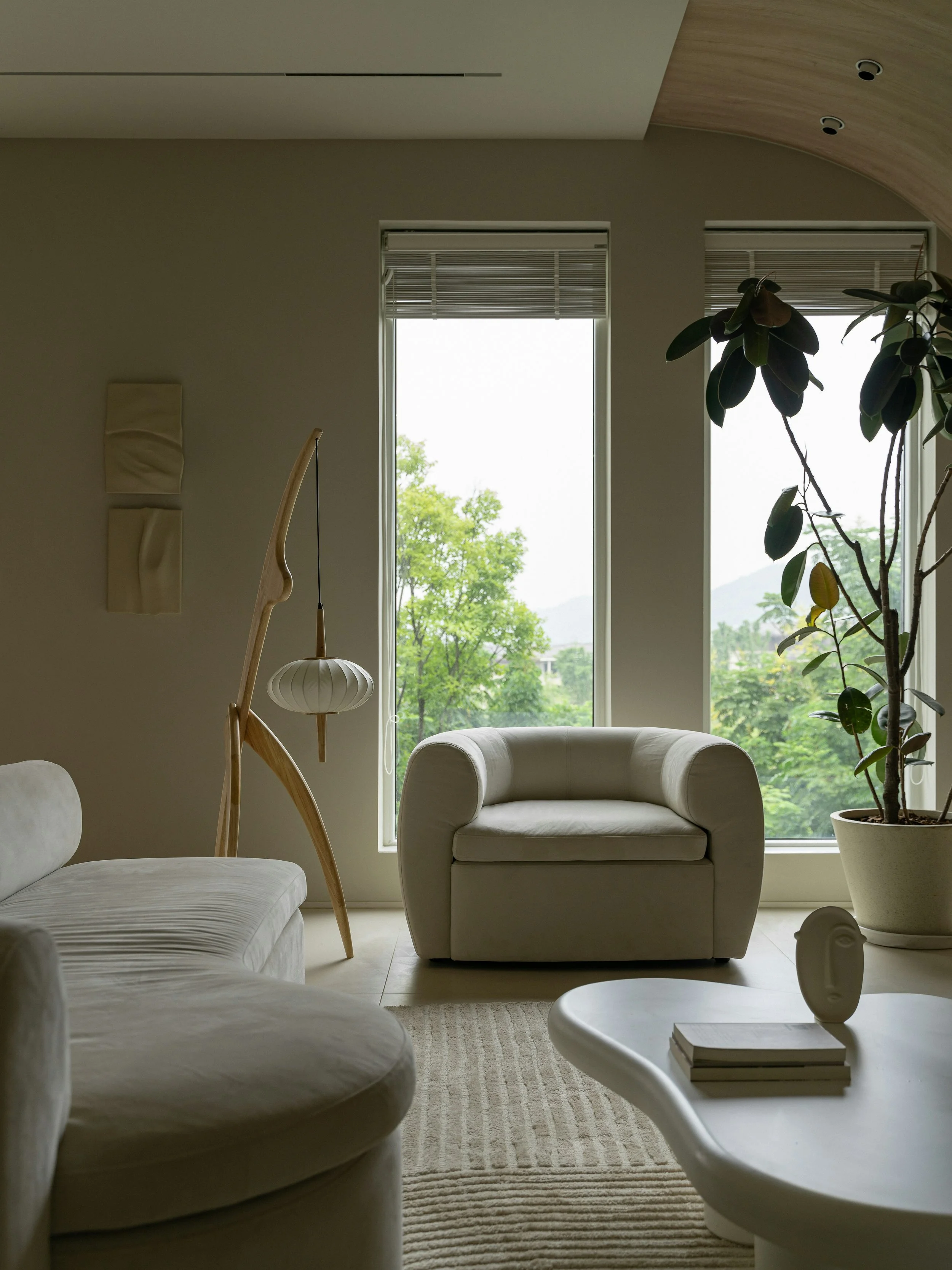 Modern living room with beige furniture, large windows with view of green trees, potted plant, and designer light fixture.