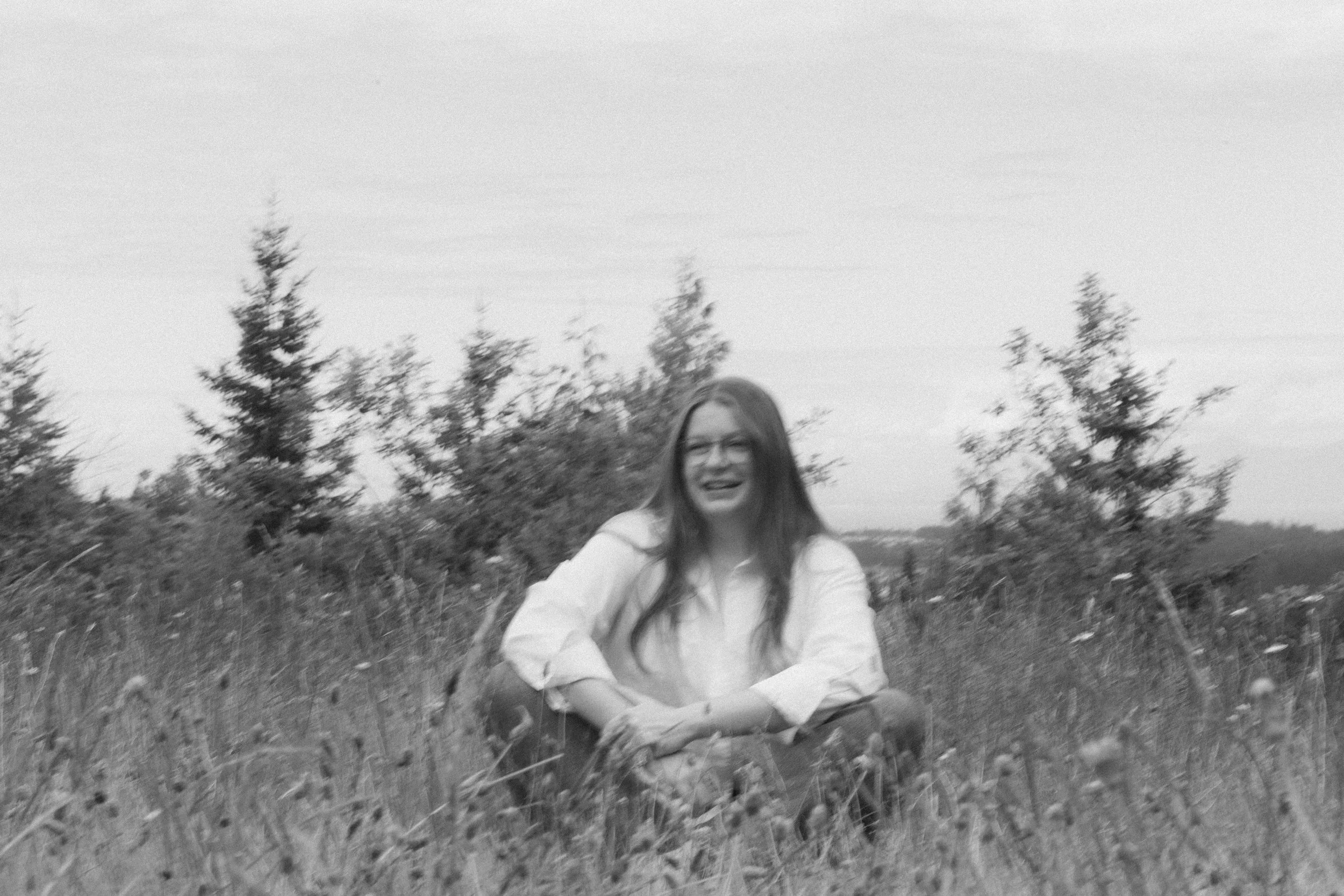 Black and white photo of a woman with long hair sitting on the ground outdoors with trees in the background, smiling and looking at the camera.