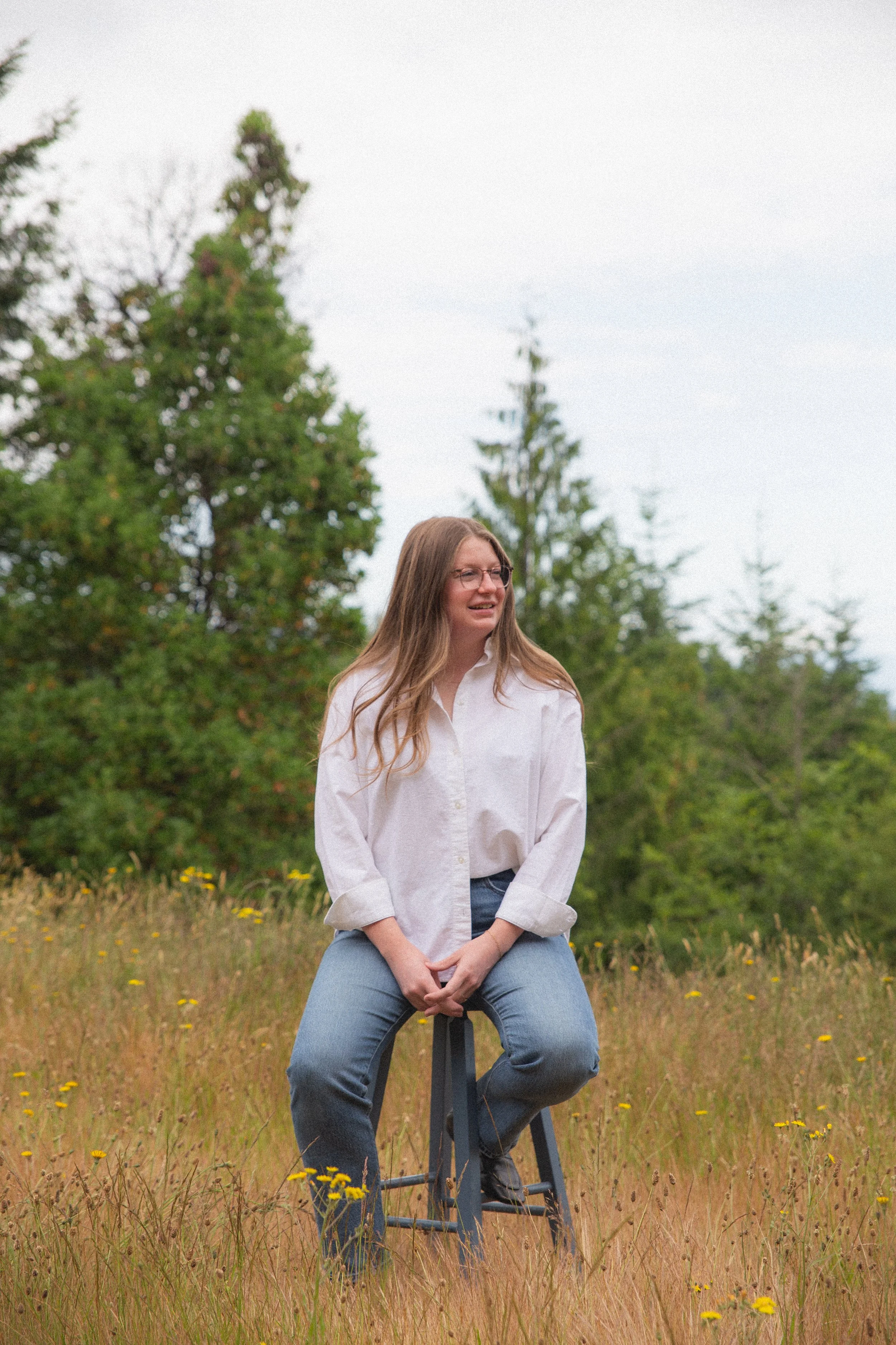 A woman with long brown hair and glasses sitting on a stool in a grassy field with yellow flowers, wearing a white shirt and blue jeans, with a background of green trees and an overcast sky.