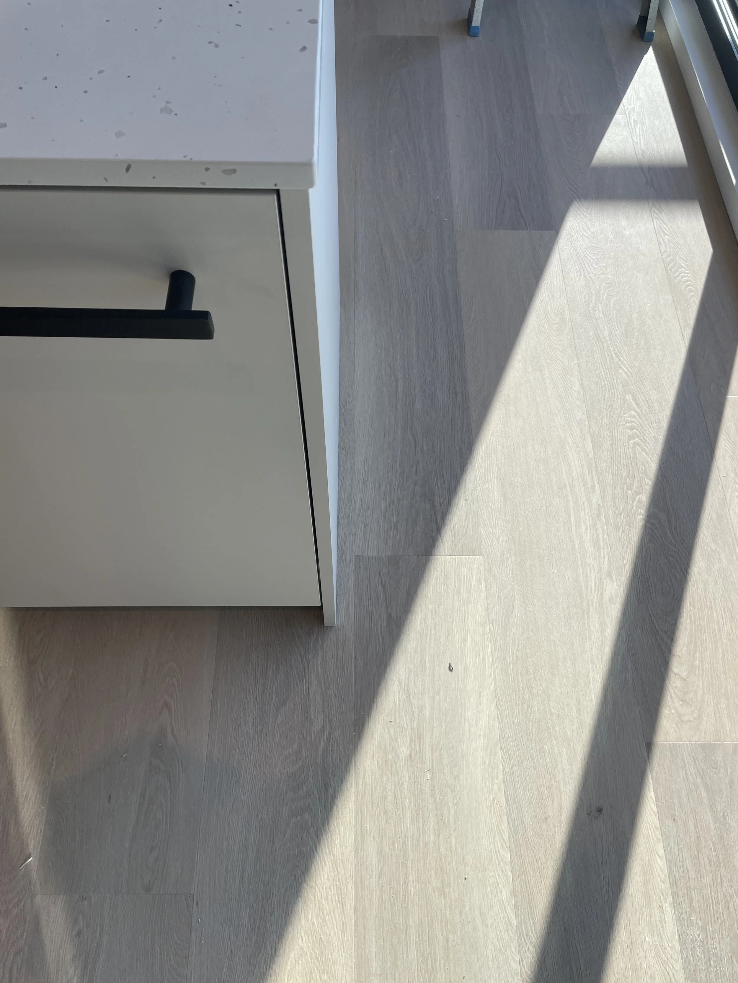 Part of a kitchen or office space showing a white cabinet with a black handle, a terrazzo-style countertop, and light-colored wood flooring with sunlight casting shadows.