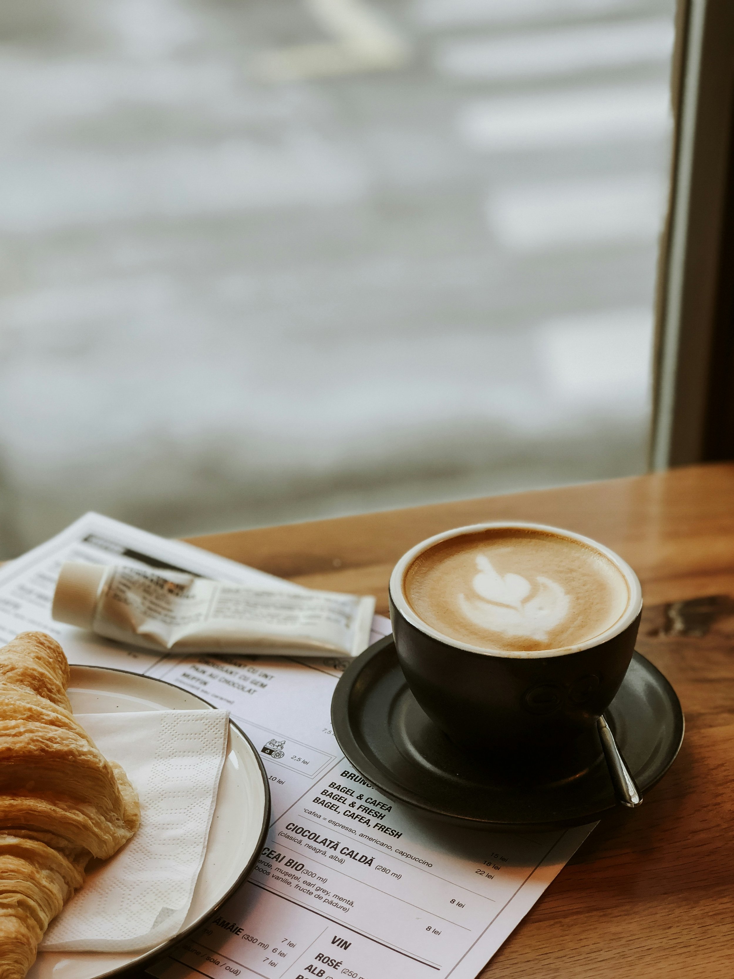 A breakfast table with a cup of coffee on a black saucer, a plate of croissants, and a newspaper on a wooden table near a window.