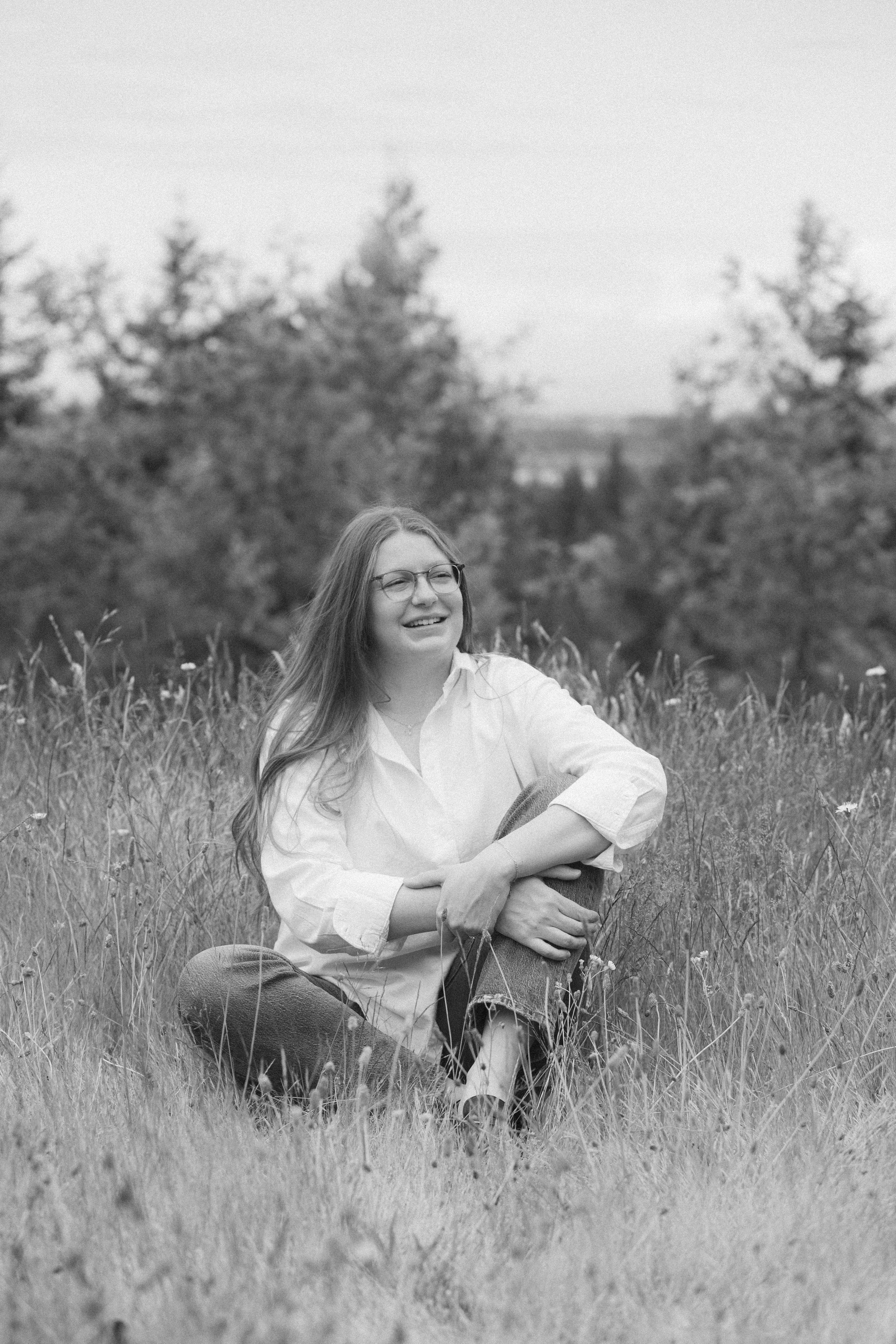 A smiling woman with glasses sitting cross-legged in a grassy field with tall grass and trees in the background.
