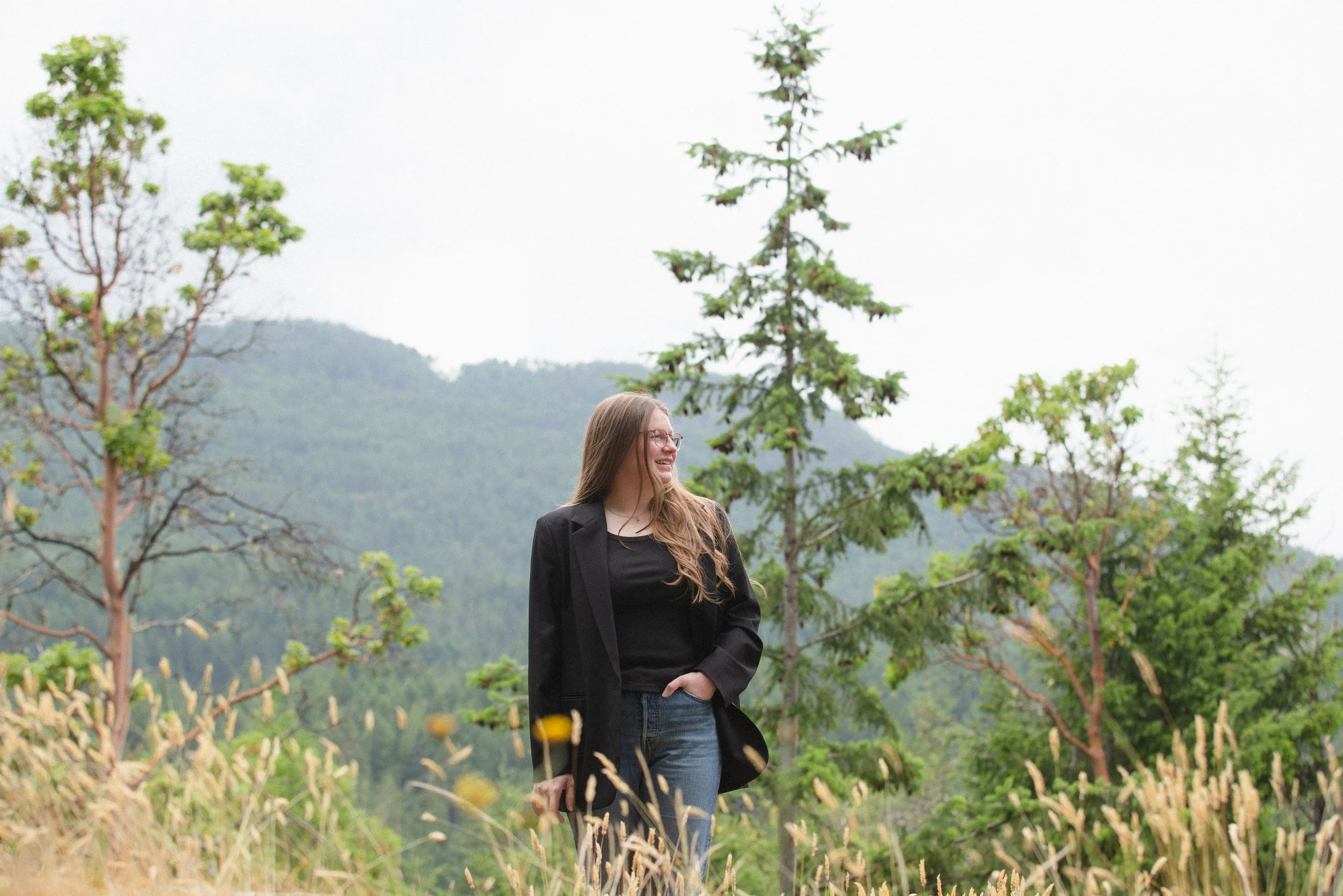 A woman with long hair, glasses, wearing a black blazer and jeans, standing outdoors among trees and mountains in the background, smiling and looking to her left.