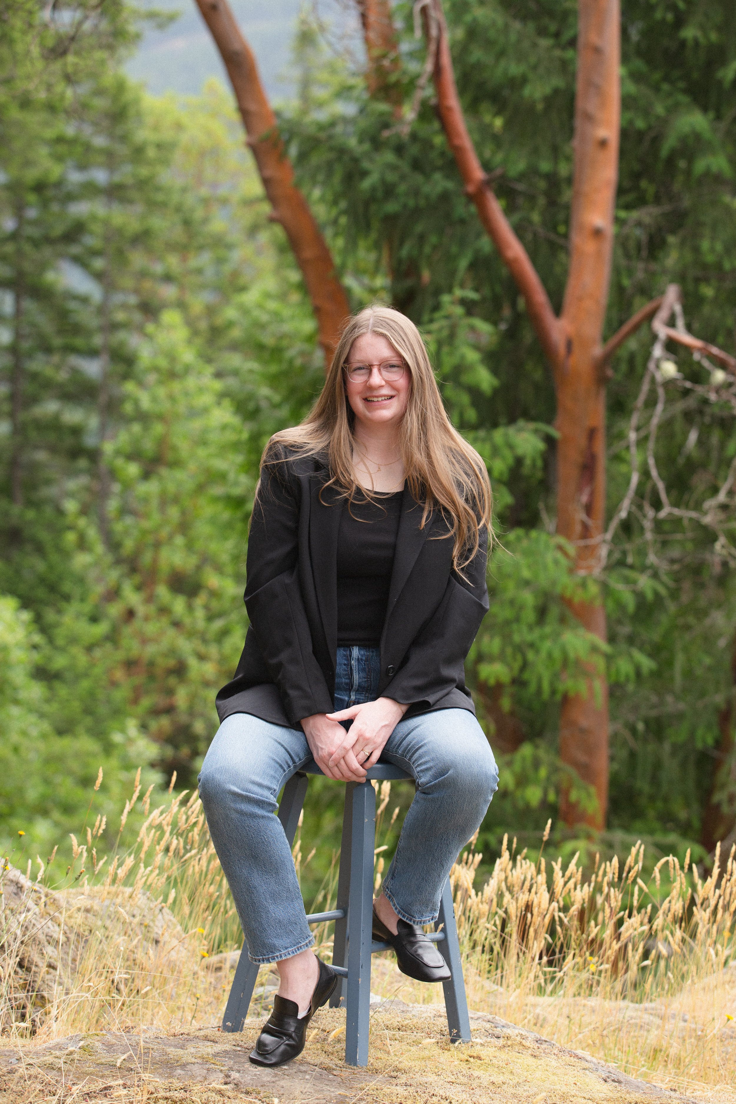 A young woman with long blonde hair, glasses, and a smile sitting on a stool outdoors in a natural setting with trees and grass.