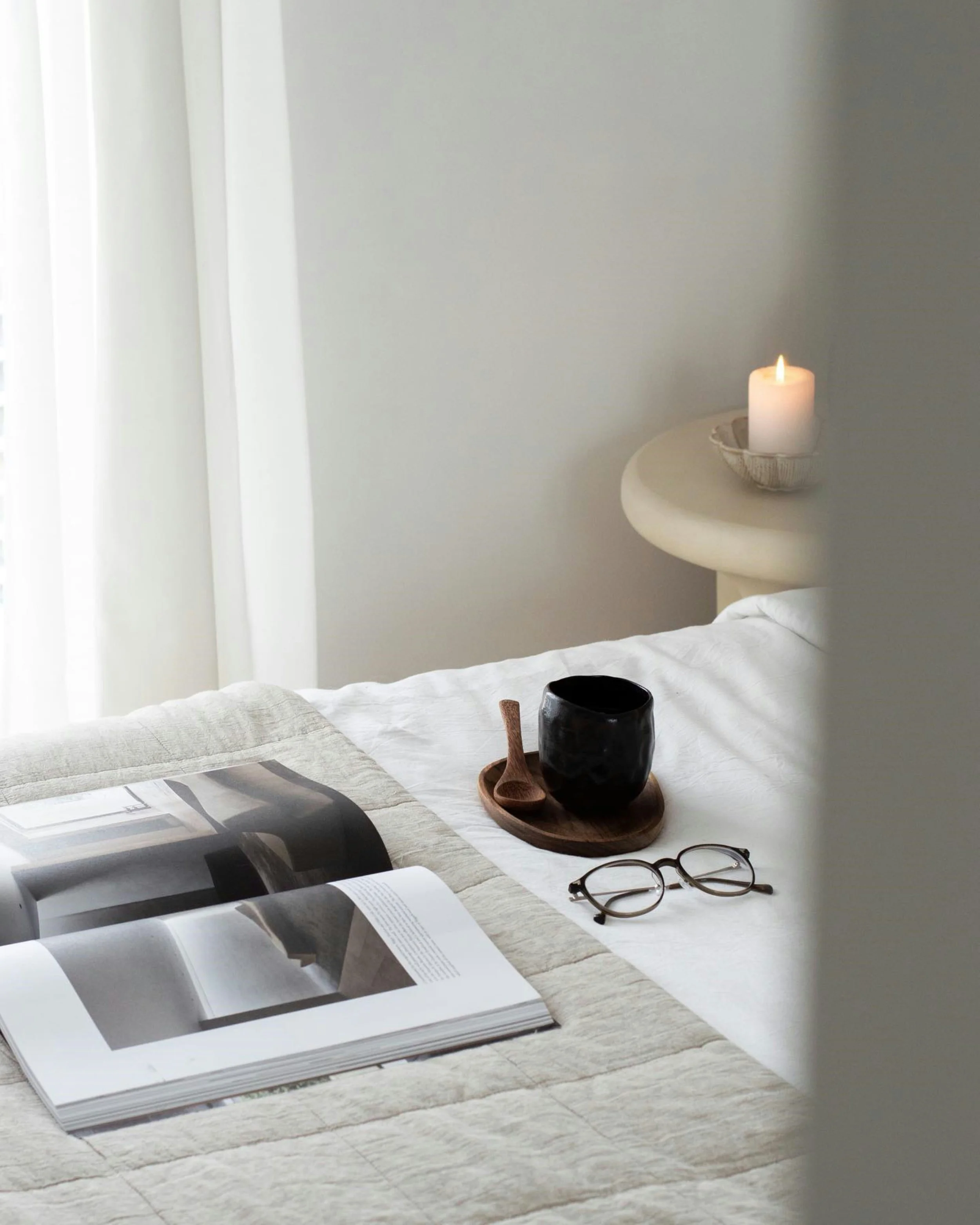 A bedroom with a white wall and a round side table that has a lit white candle in a bowl. On the bed, there is an open magazine with black and white images, a pair of glasses, and a black cup with a wooden spoon on a round wooden tray.