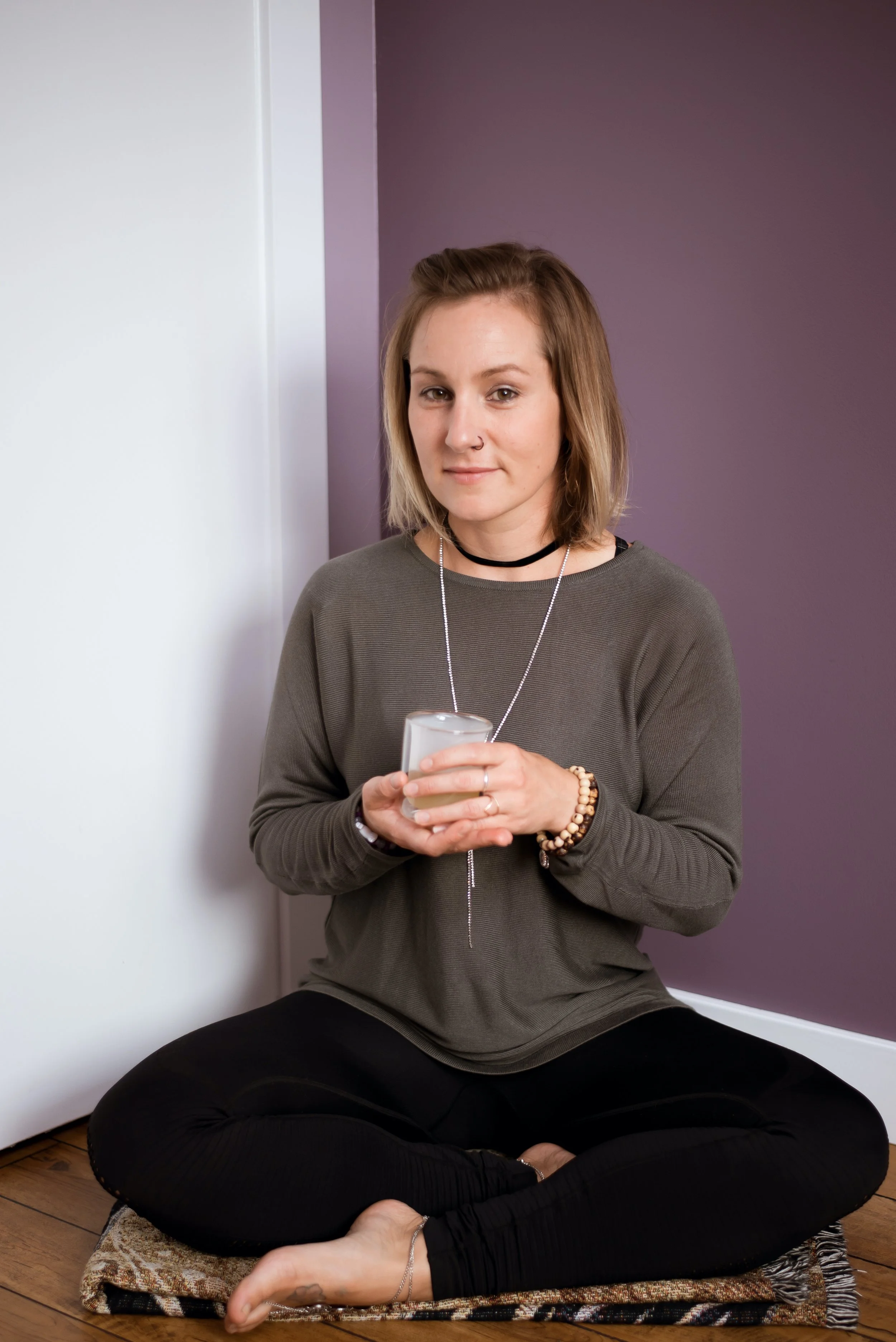 A young woman sitting cross-legged on a small rug on the floor, holding a candle in her hands, with a calm expression.