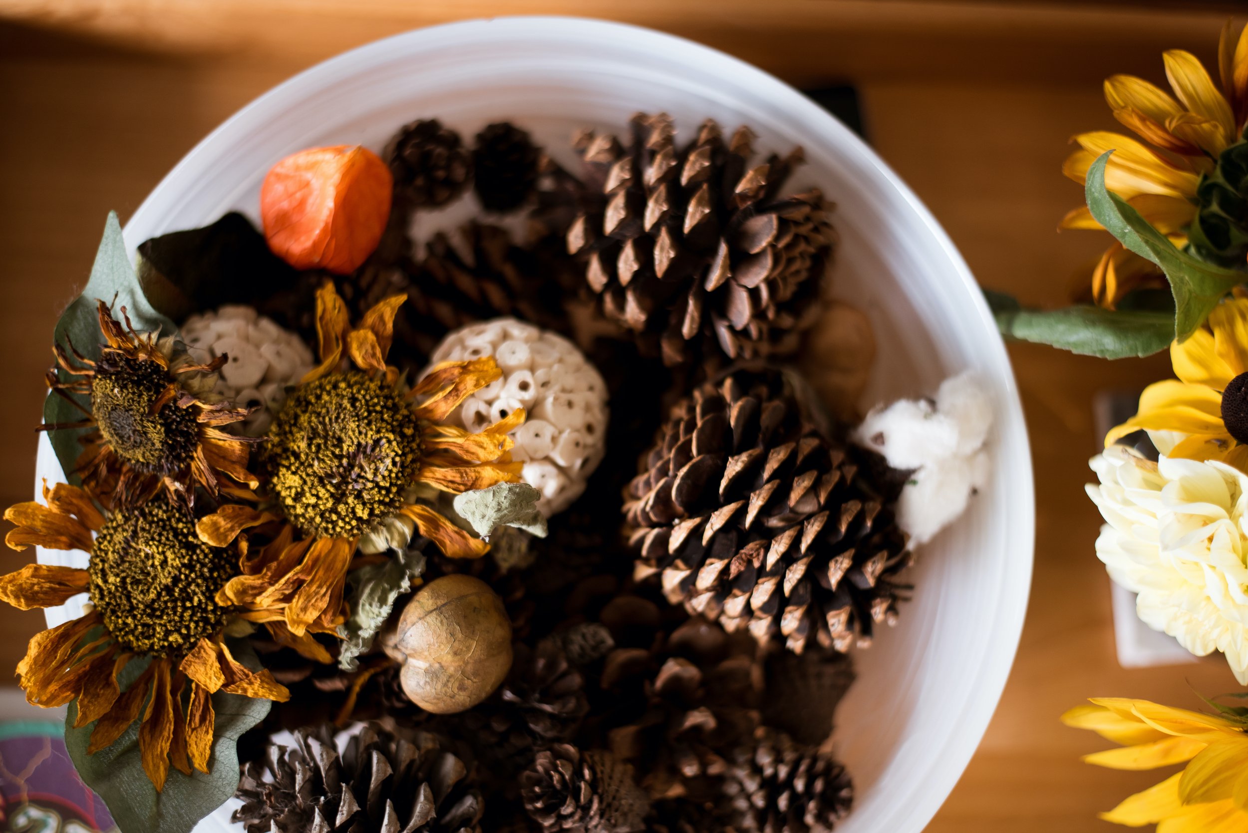 A white bowl filled with pinecones, dried flowers, and a small orange pumpkin, with part of a bouquet of yellow flowers visible on the right.