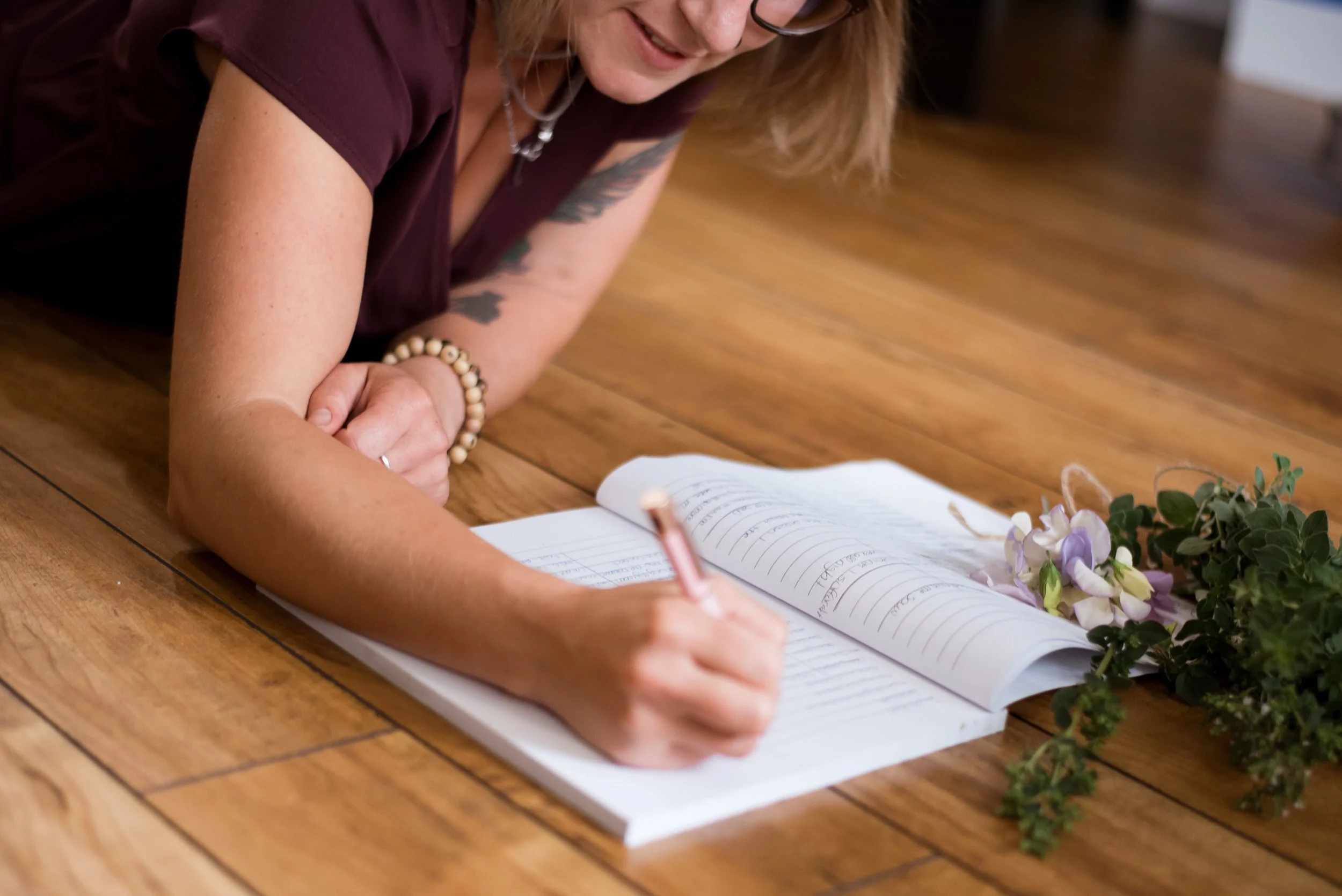 A woman lying on a wooden floor and signing a guestbook with a pen, with flowers nearby, wearing a burgundy shirt, glasses, bracelet, and a necklace.