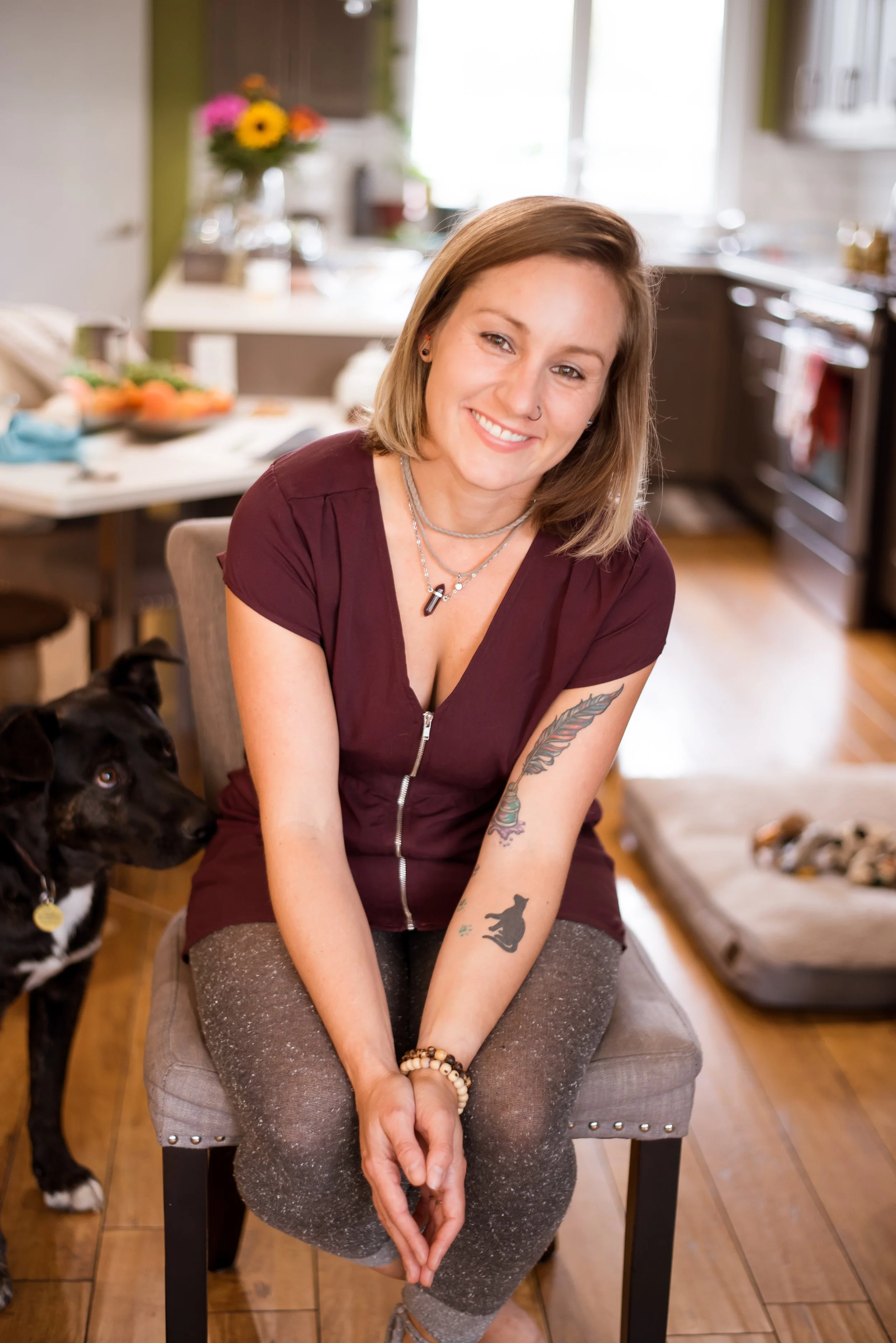 A young woman with shoulder-length light brown hair, wearing a maroon zip-up top, gray sparkly pants, and jewelry including necklaces and bracelets, sits on a beige upholstered chair in a kitchen. She is smiling at the camera with tattoos on her left arm, and a black and white dog stands next to her looking up. The background shows a kitchen counter with flowers, food, and various items.
