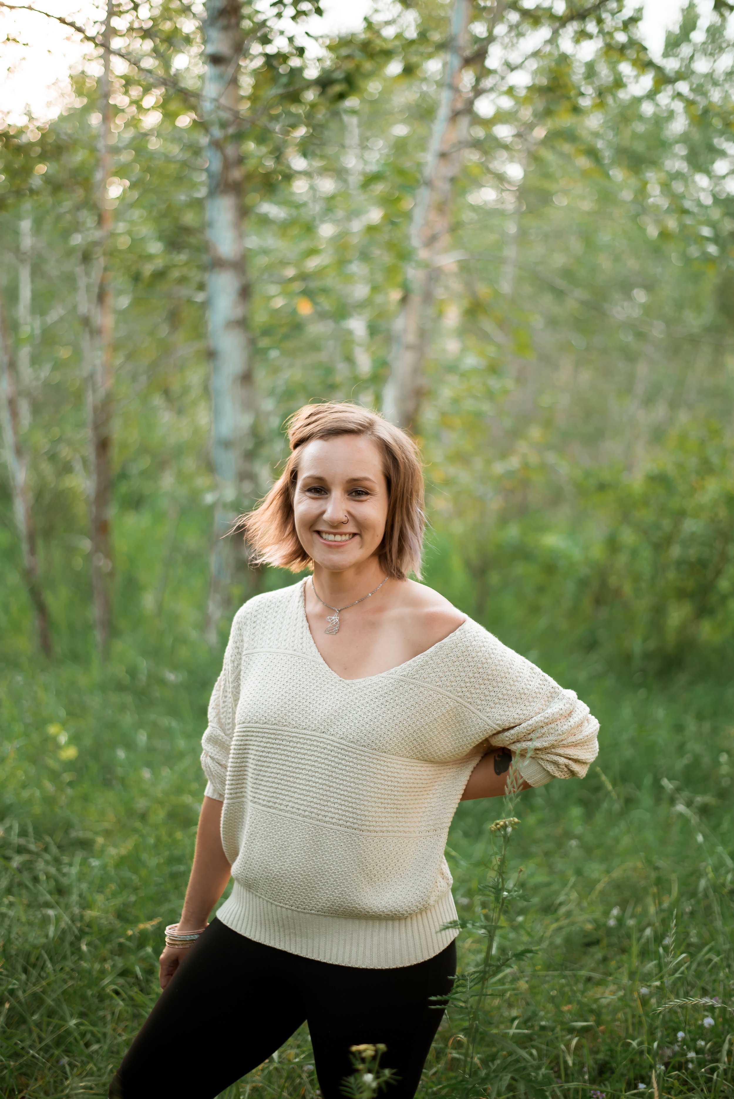 A smiling woman with shoulder-length light brown hair, wearing a cream-colored knitted sweater and black pants, standing outdoors in a green, wooded area during daylight.