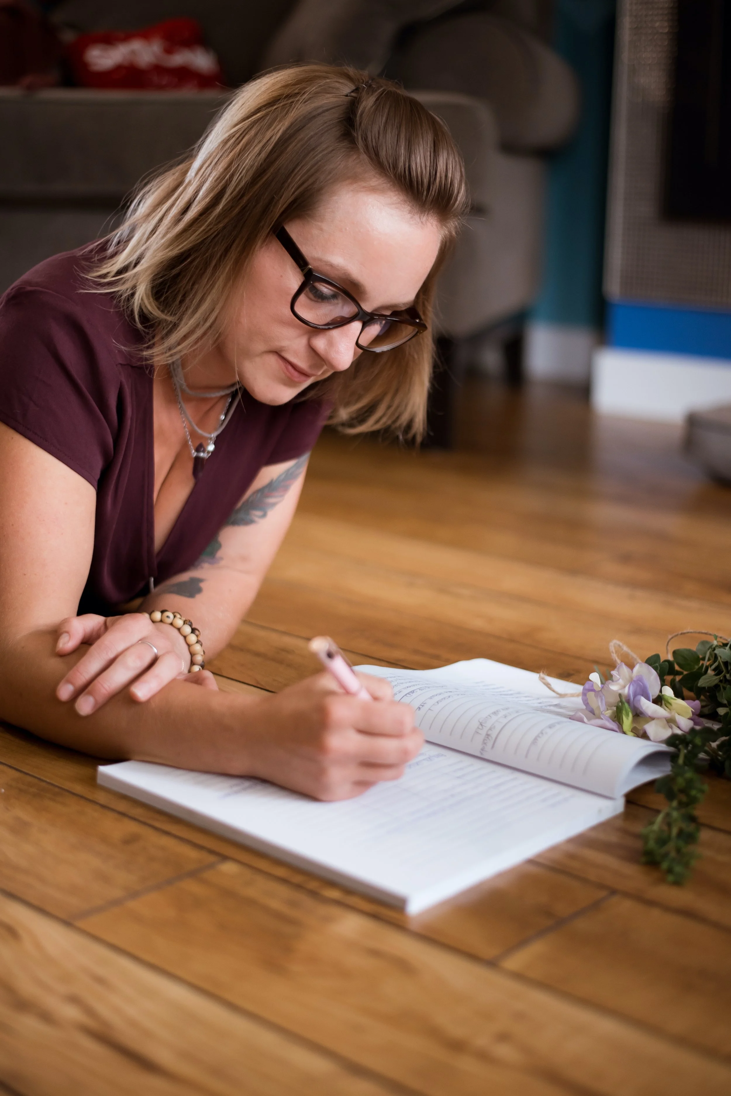 A woman with glasses and tattoos lies on the wooden floor, writing in a book with a pink pen. There are flowers beside her.