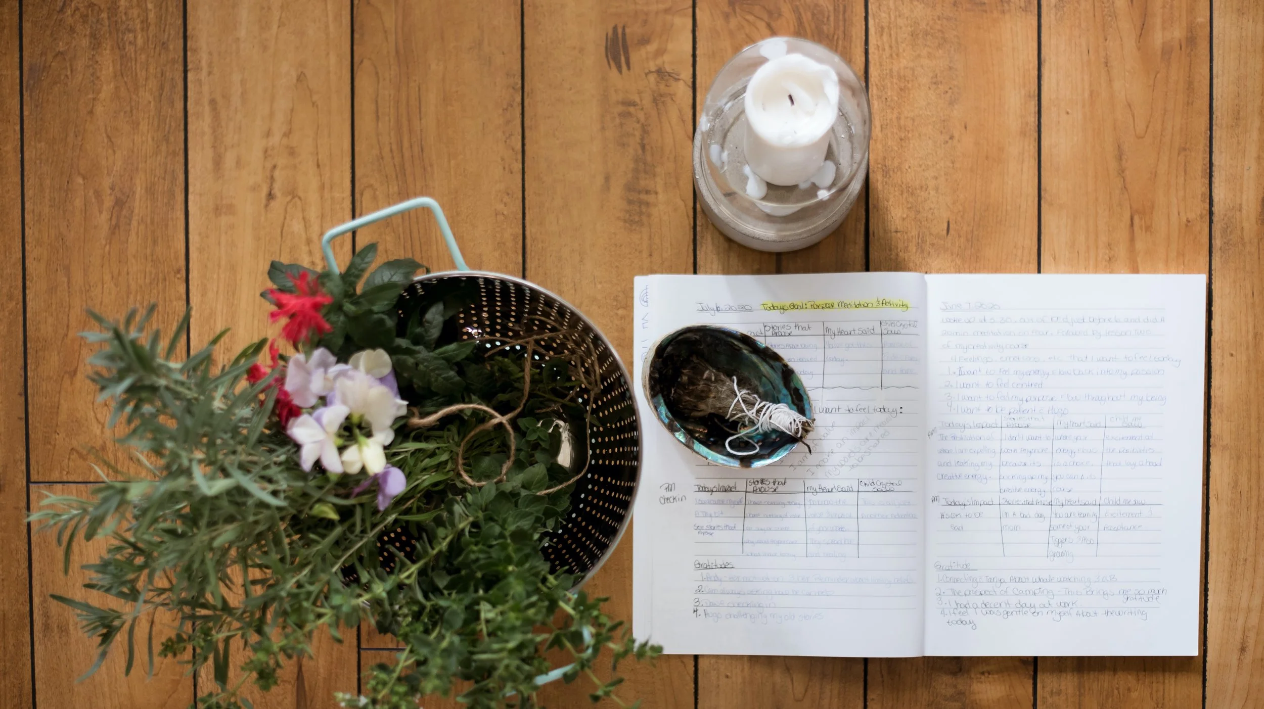 Top-down view of a wooden table with a potted plant, a notebook with handwritten notes, a small bowl with a tied bundle of herbs, a lit white candle in a glass holder, and a glass container with water and white candle wax.