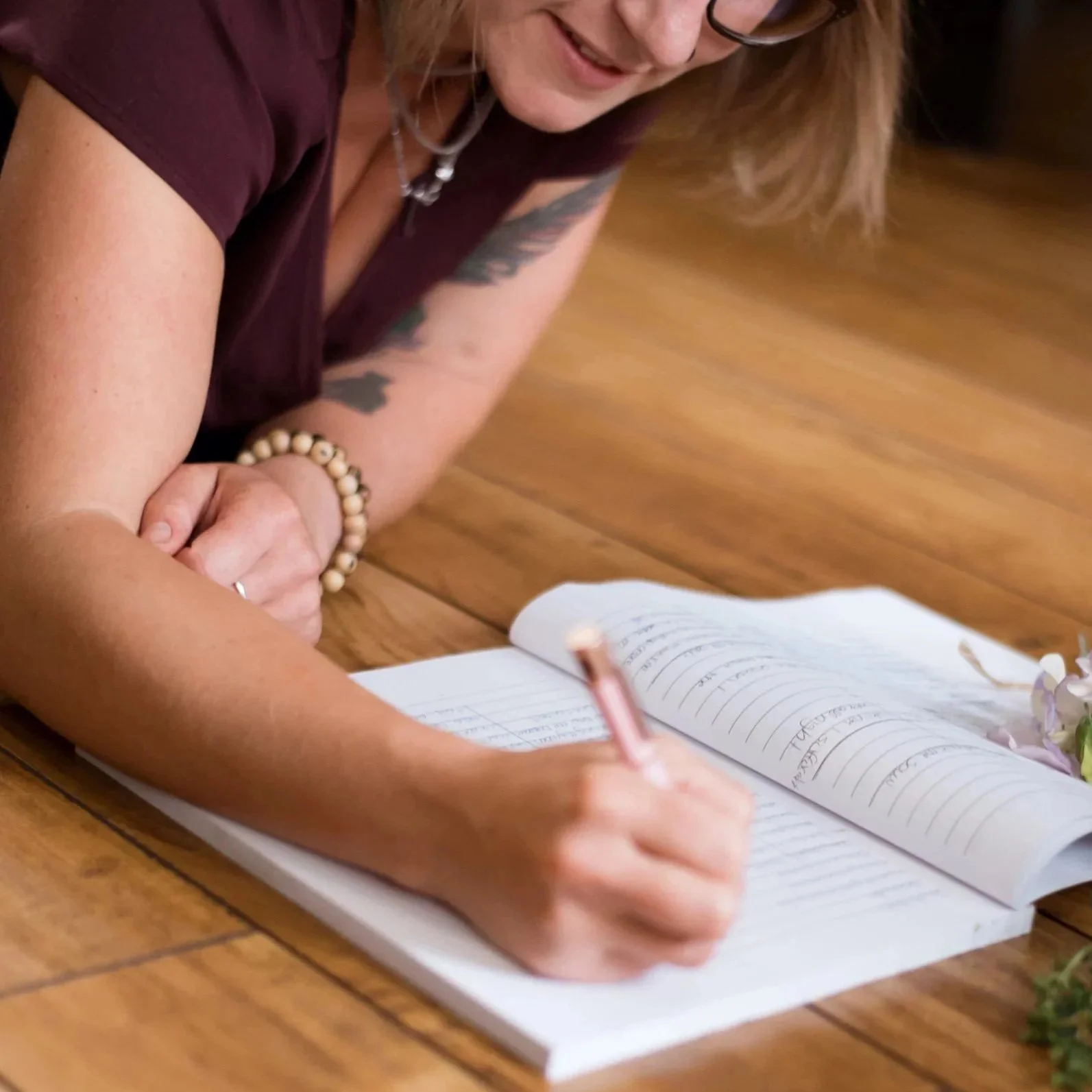 A woman with glasses and tattoos is lying on the wooden floor, signing a guestbook with a pen.