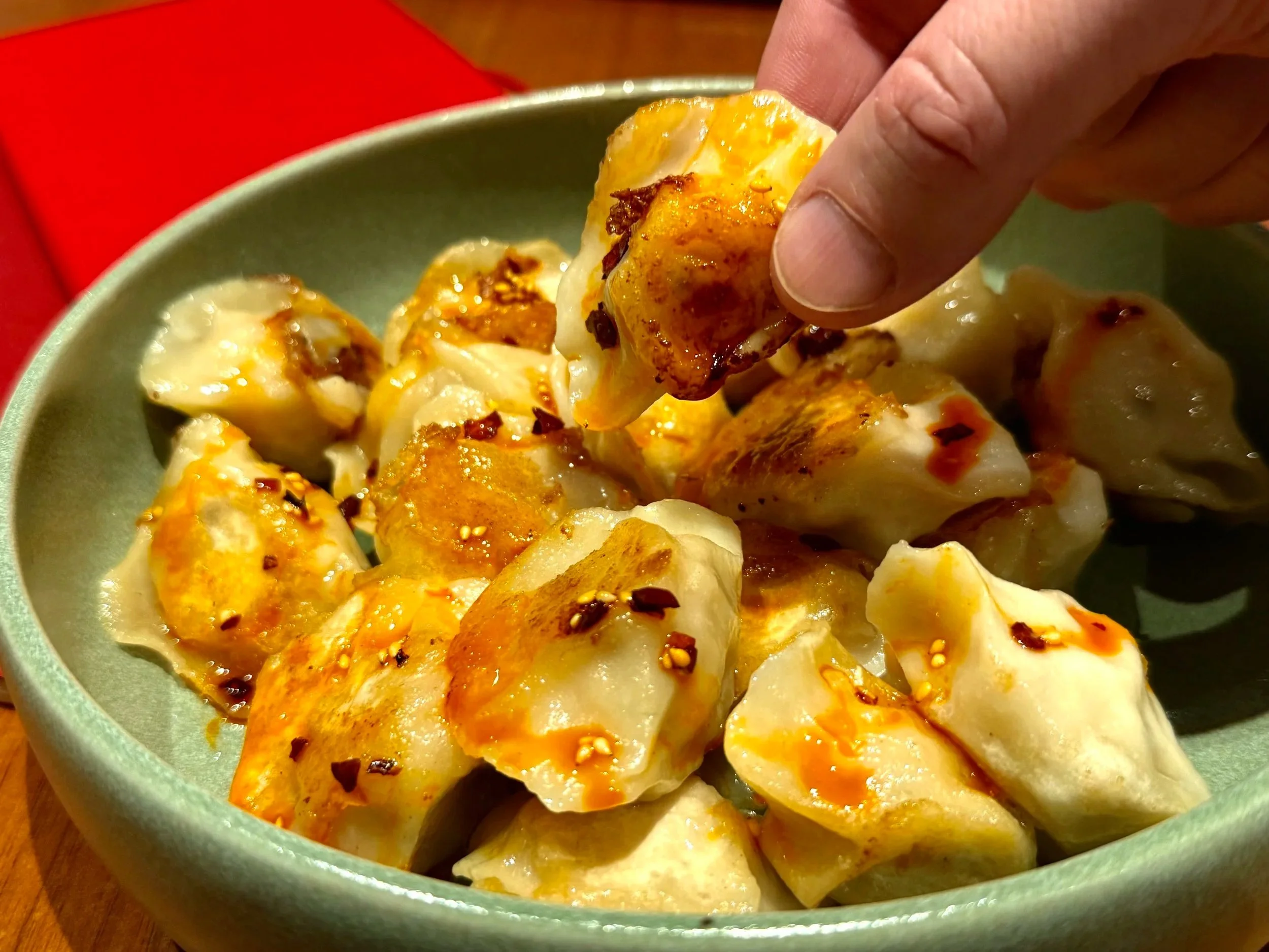 A close-up of a bowl filled with cooked dumplings, with a person's hand lifting one dumpling, showing the golden-brown, crispy surface and small chili flakes on top.