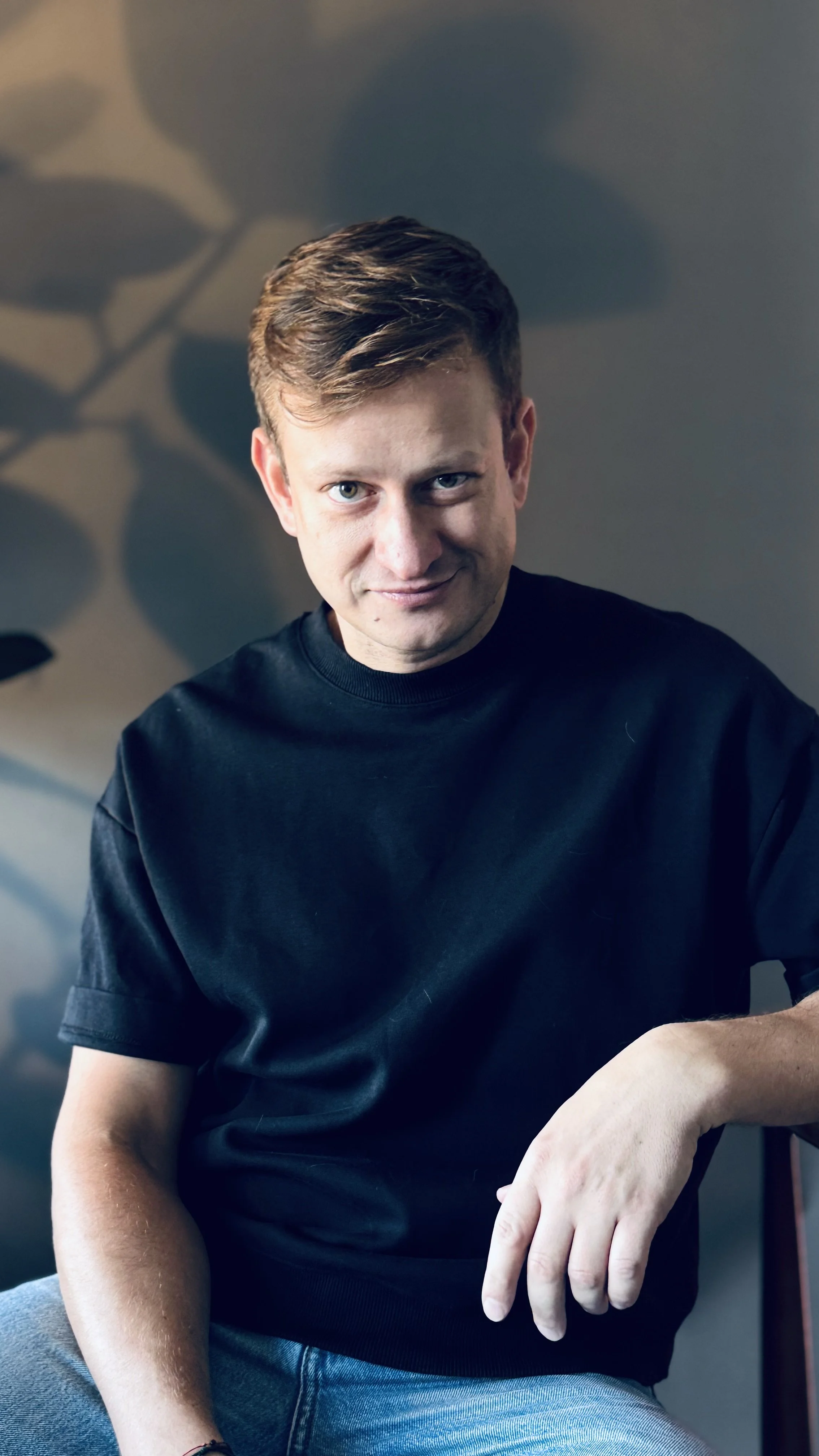 A young man with light brown hair, wearing a black T-shirt, sitting indoors against a background with shadows of leaves on the wall, smiling slightly at the camera.