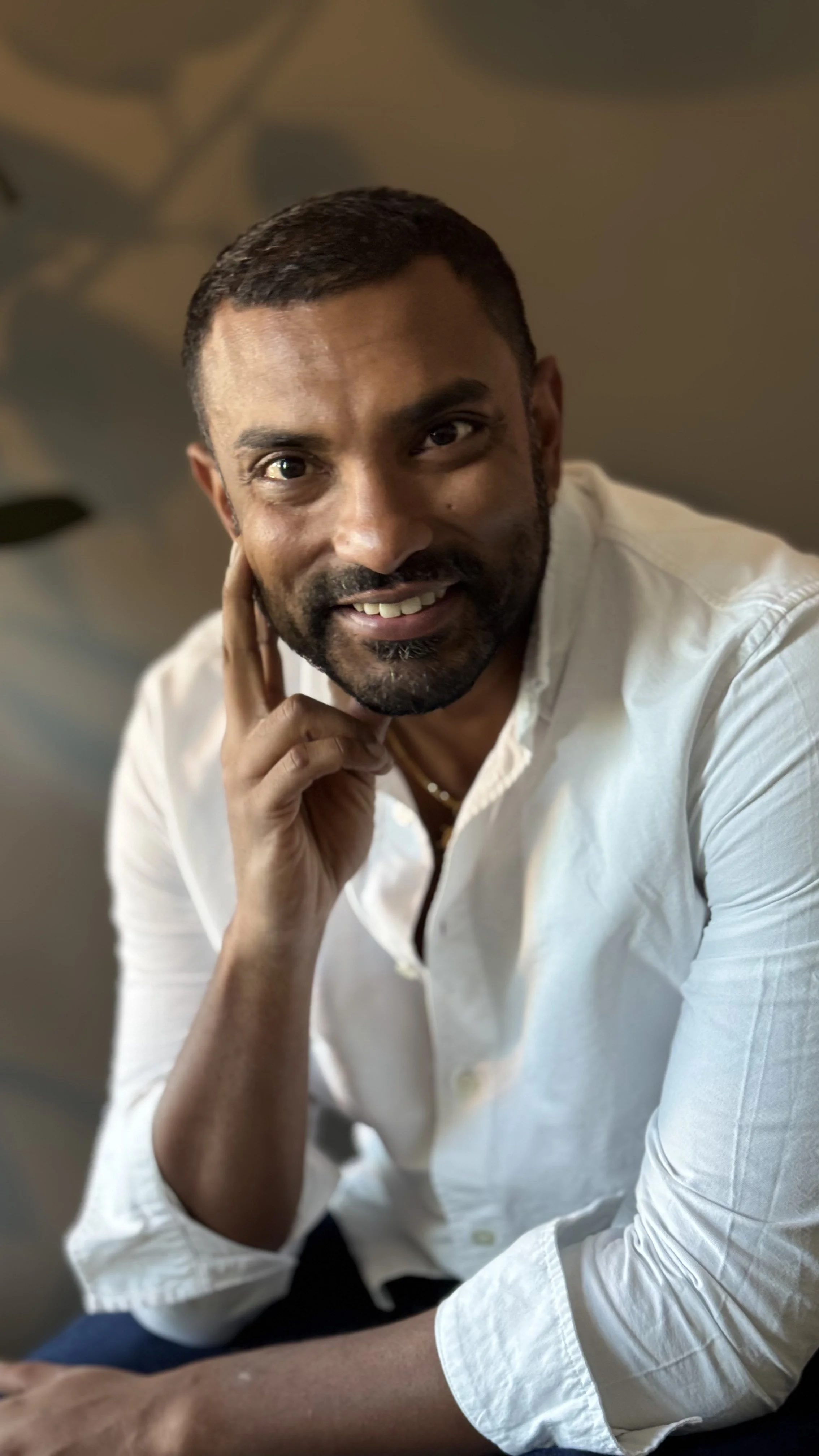 A man with short dark hair and a beard, wearing a white shirt, smiling and resting his hand on his face.