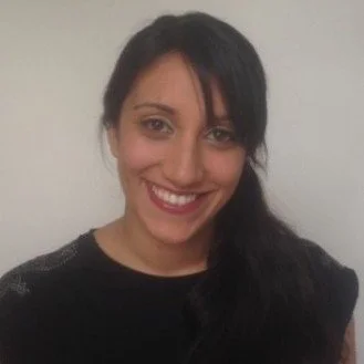 A woman with dark hair and a bright smile, wearing a black top, standing against a plain light-colored background.