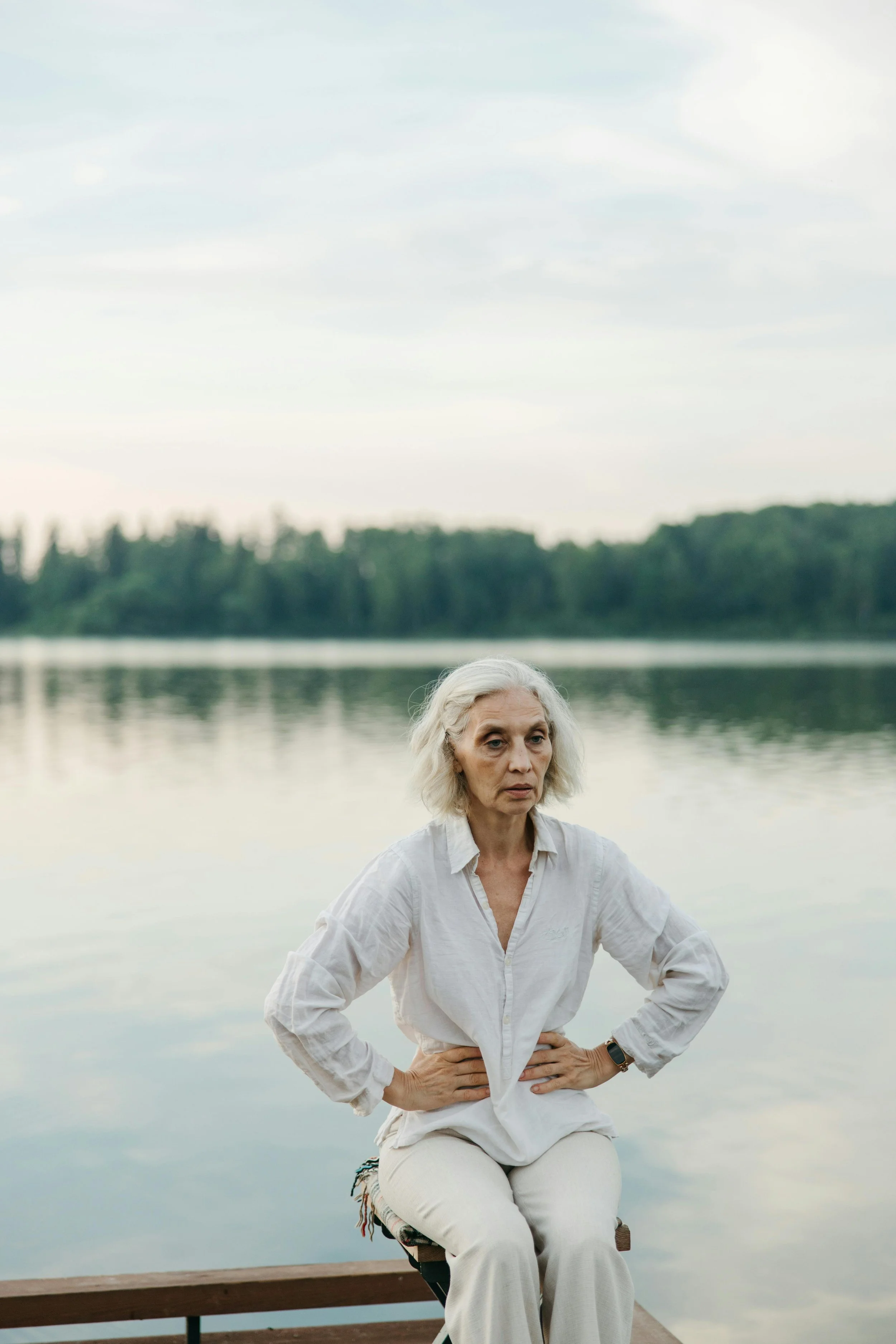 Older woman sitting by a calm lake with hands on hips, appearing reflective and emotionally heavy in a peaceful natural setting.