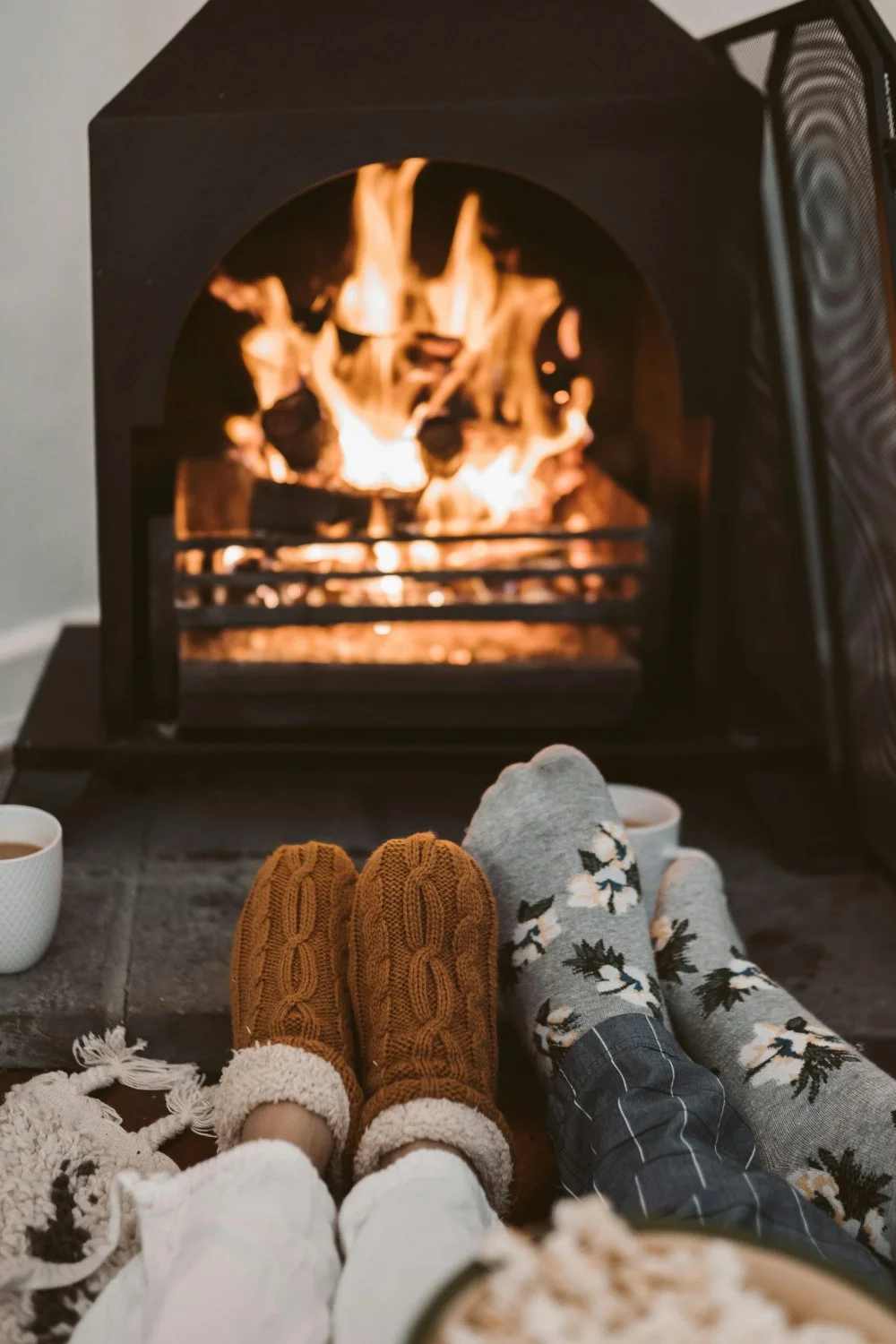 Two people sitting by a lit fireplace with warm socks and slippers, cups of hot drinks nearby, creating a cozy winter scene.