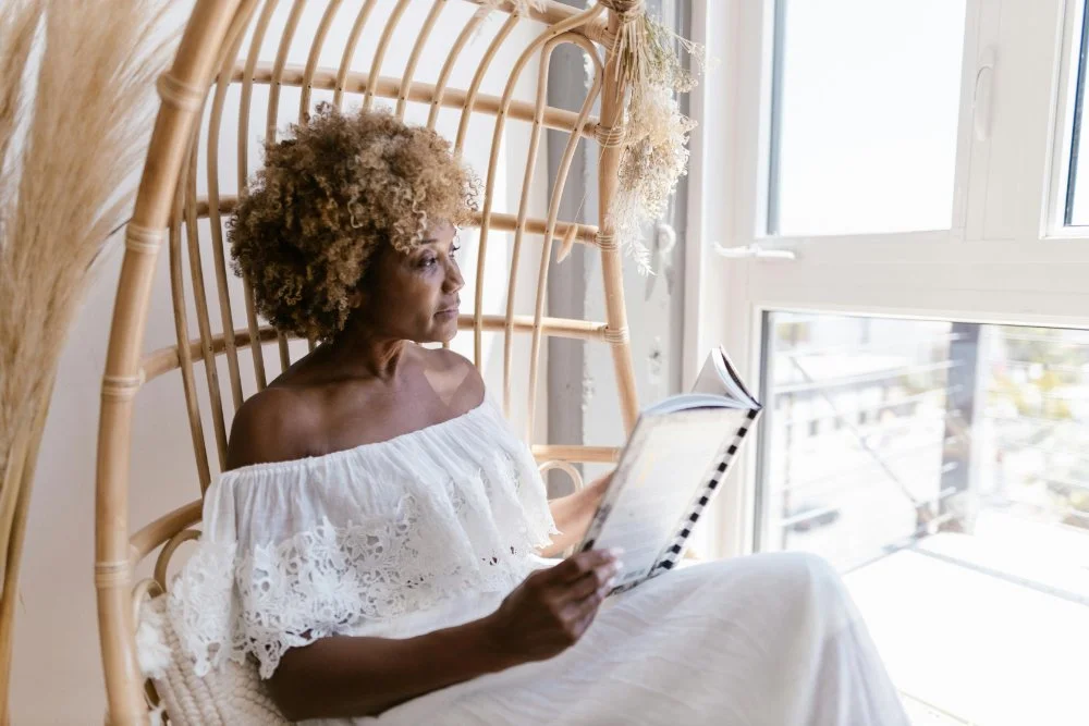 Woman reading in a bright, cozy room, seated in a woven chair by the window. | EMDR therapy NYC