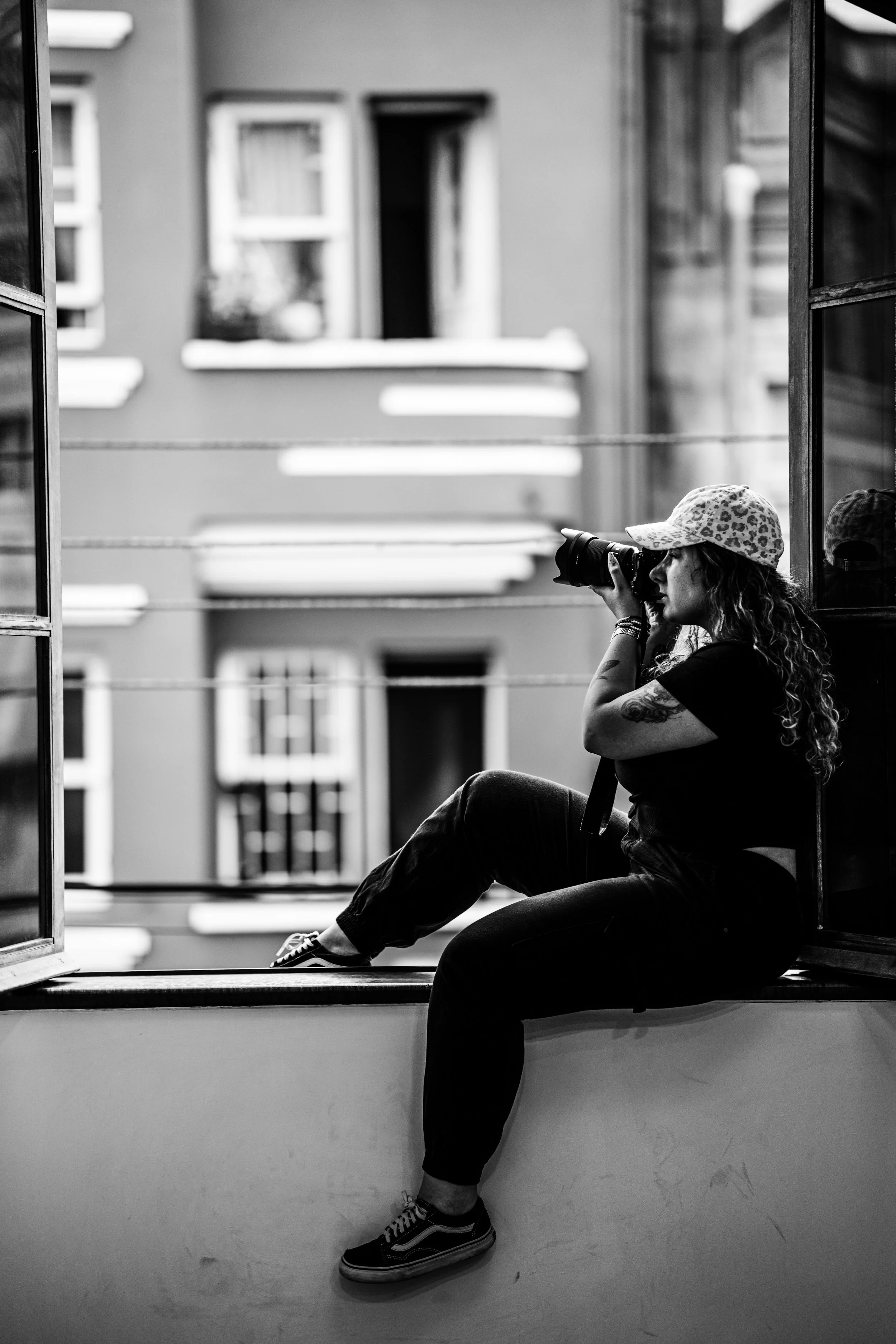 a black and white photo of a woman sitting outside a window taking a photo with a professional camera.