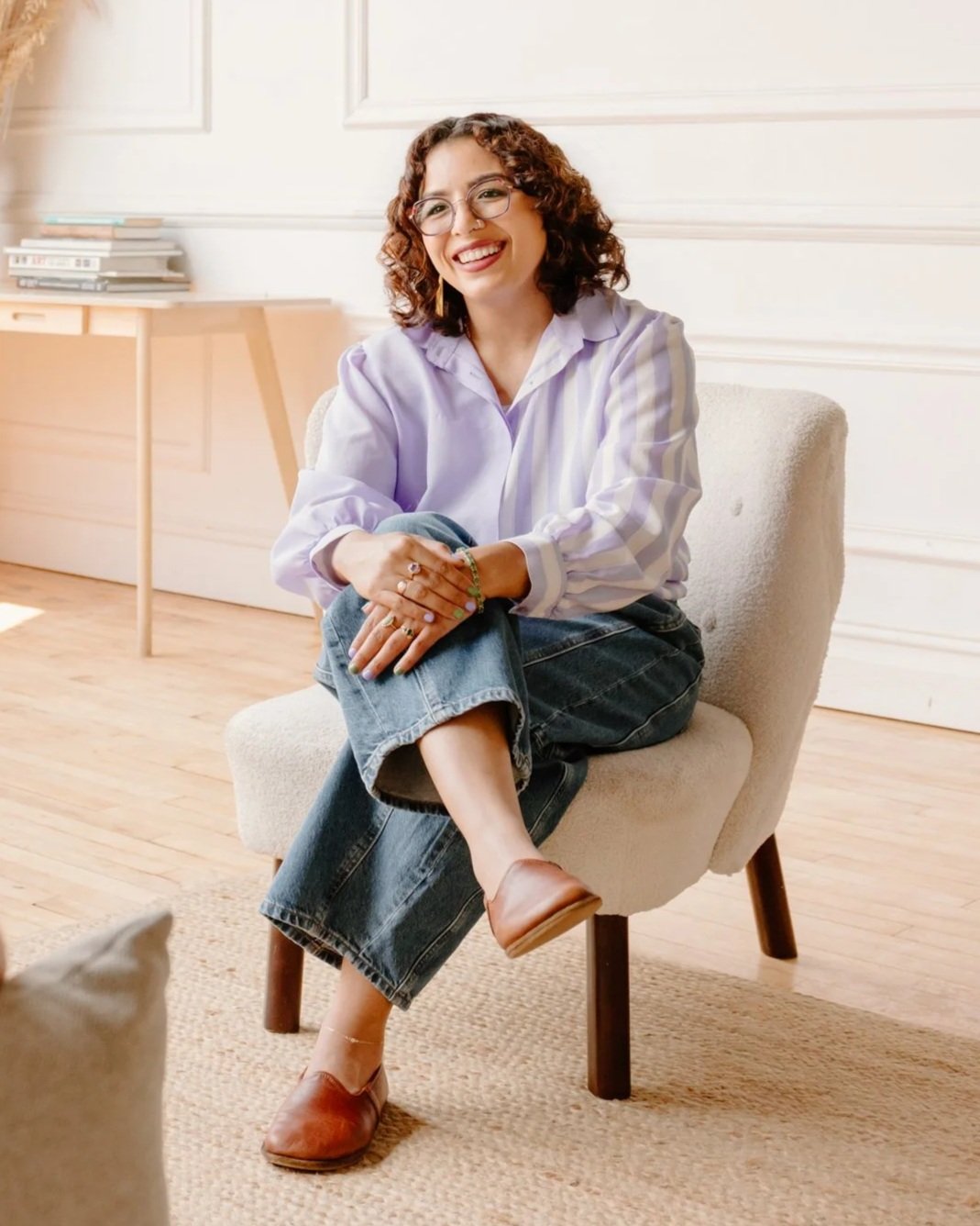EMDR and DBR therapist Jennifer Budhan seated in a bright, welcoming office, smiling warmly with legs crossed as she listens attentively to her client.