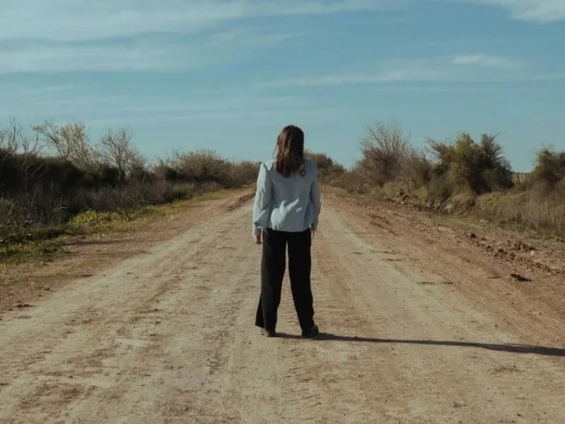 Person stands alone on a wide dirt road, seen from behind, facing an open landscape under a large sky, with sparse trees and a sense of distance. | i dont know who i am anymore