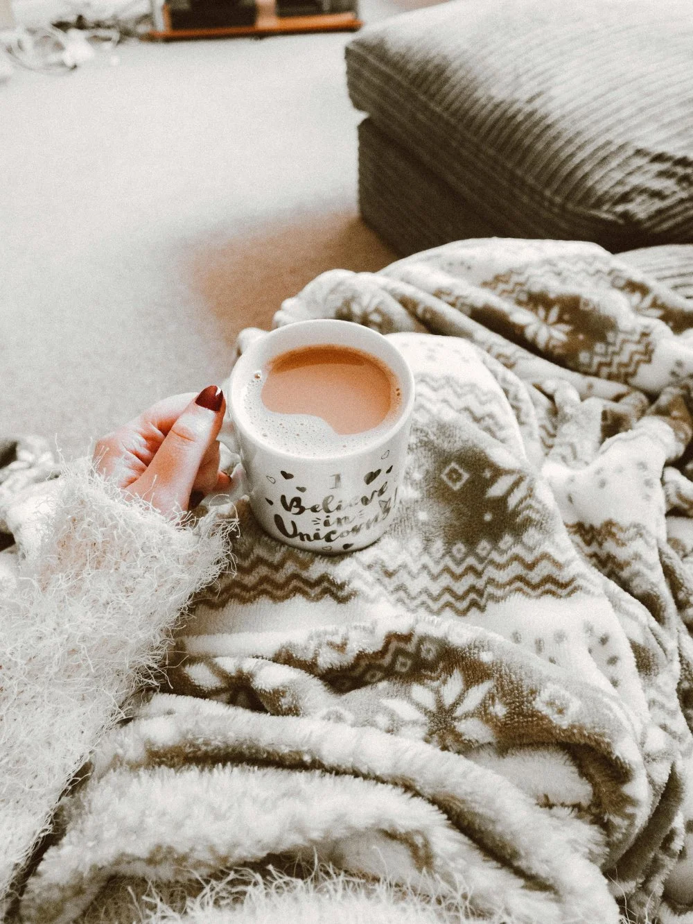 A person holding a warm drink while wrapped in a soft patterned blanket, sitting on a cozy couch in a calm, carpeted living room.