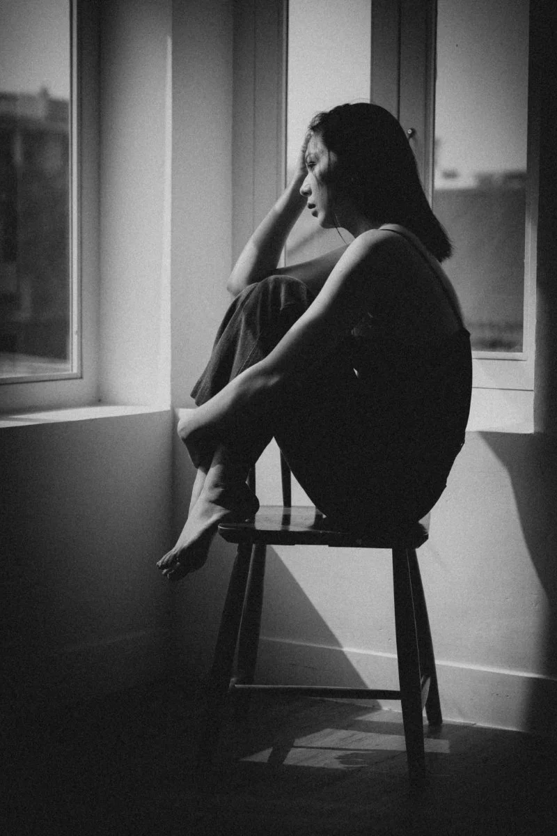 Black-and-white photo of a person sitting sideways on a stool by a window, knees drawn up, head resting on hand, light casting strong shadows in a quiet interior.