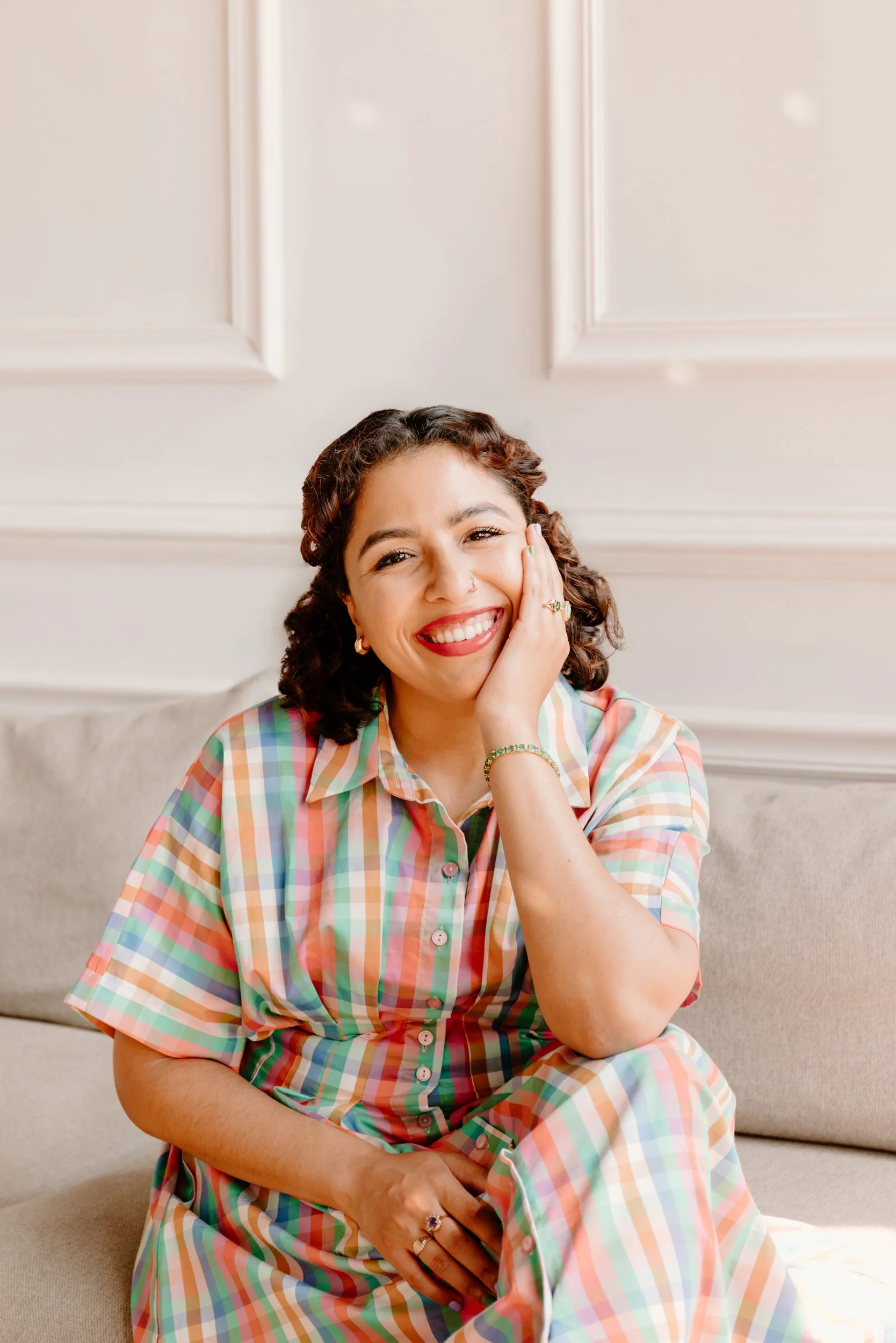 EMDR and DBR therapist Jennifer Budhan seated in a bright, welcoming office, smiling warmly with a hand resting on her cheek.