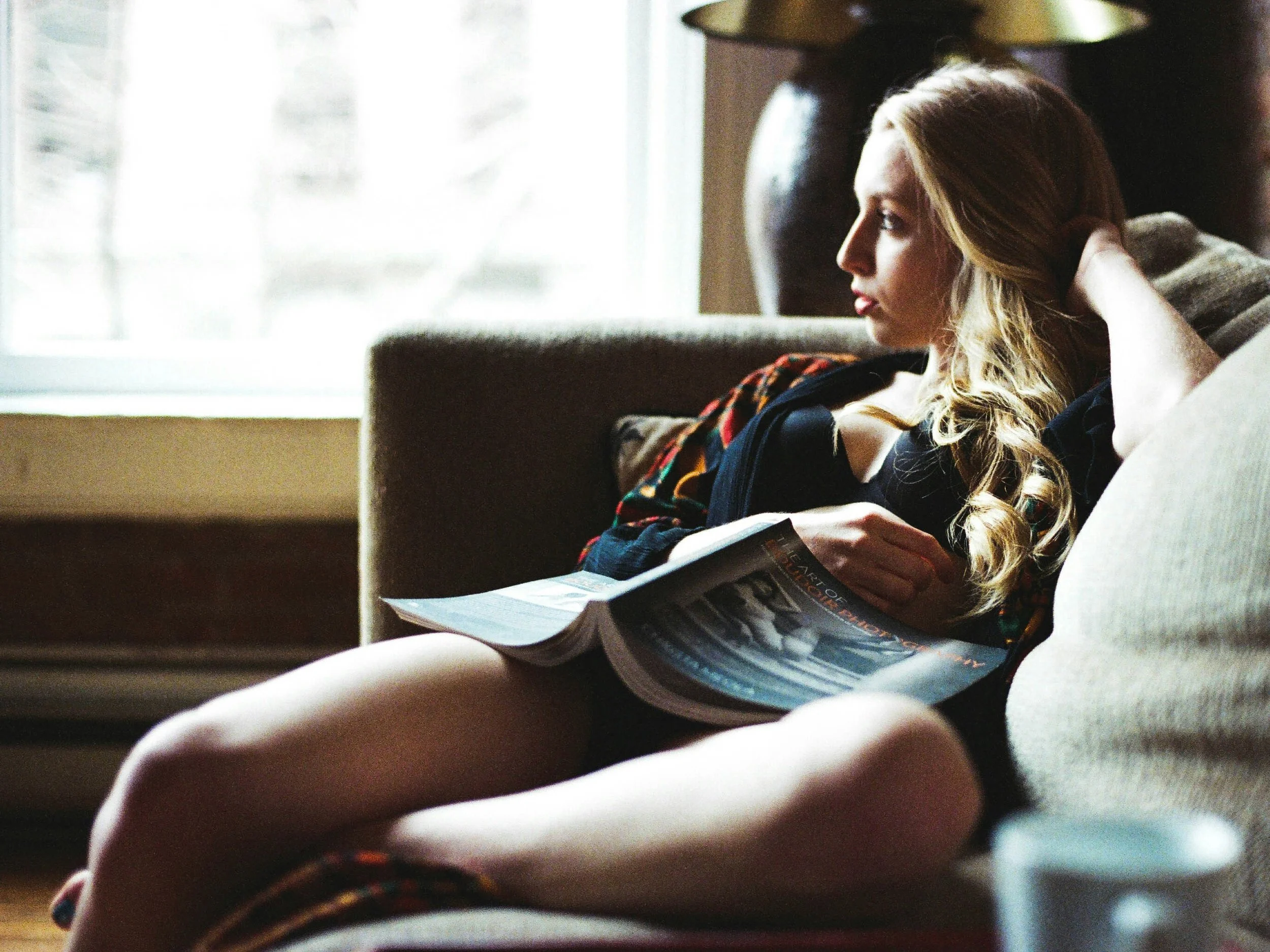 [Image: A woman reclines on a couch holding an open magazine, gazing out a window with a thoughtful expression in soft natural light, suggesting quiet rest and reflection. | relaxation-induced anxiety